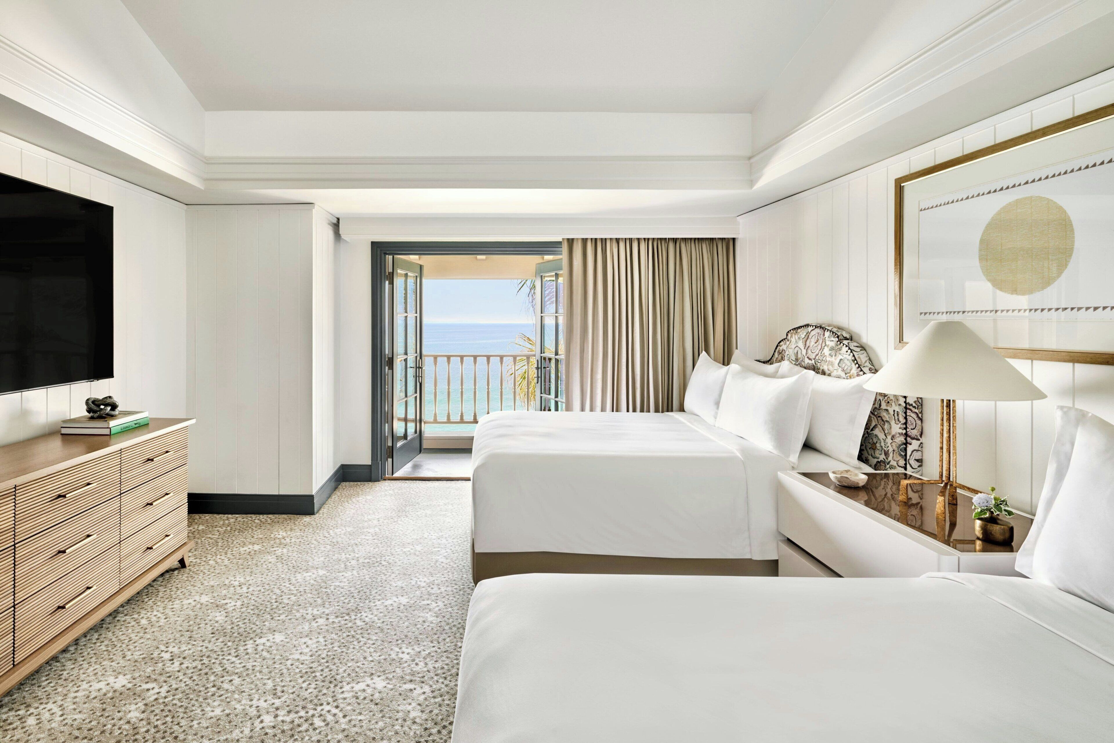 A hotel room with two beds, a TV, a wooden dresser, a bedside table with a lamp, and a view of the sea through an open door.