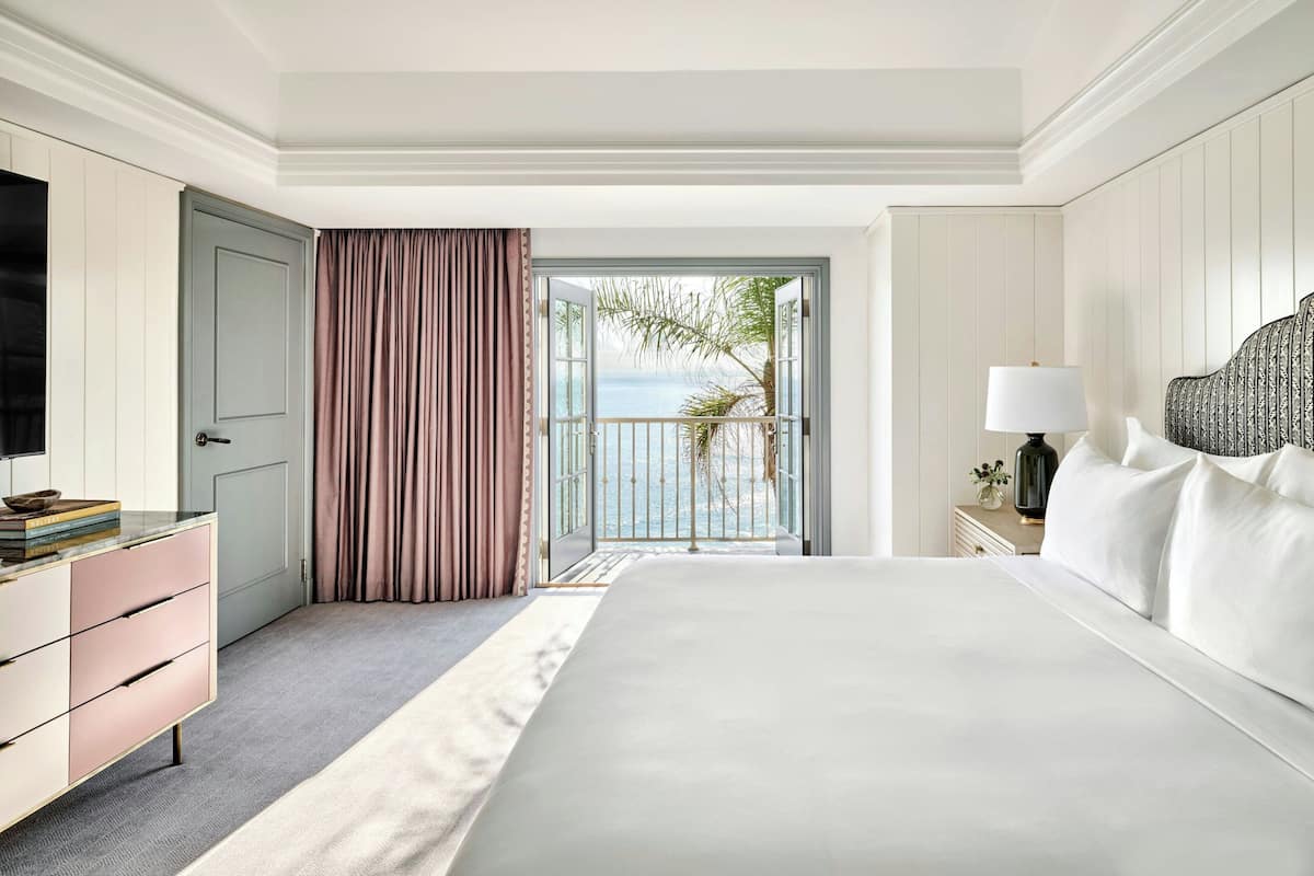A hotel room with a large bed, a television, a dresser, and a view of the sea through an open door.
