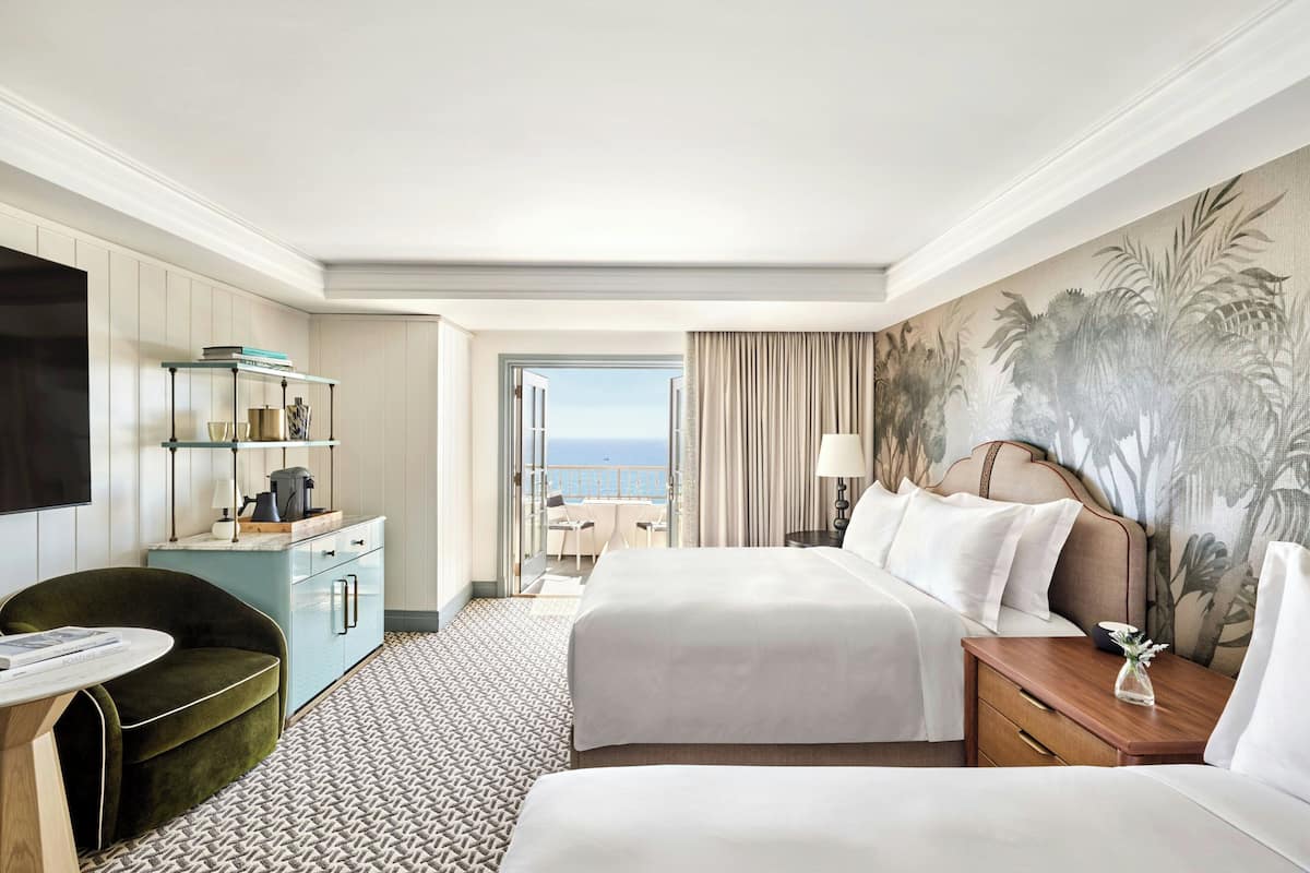 A hotel room with two beds, a desk with a chair, a TV, a minibar, and a view of the ocean.