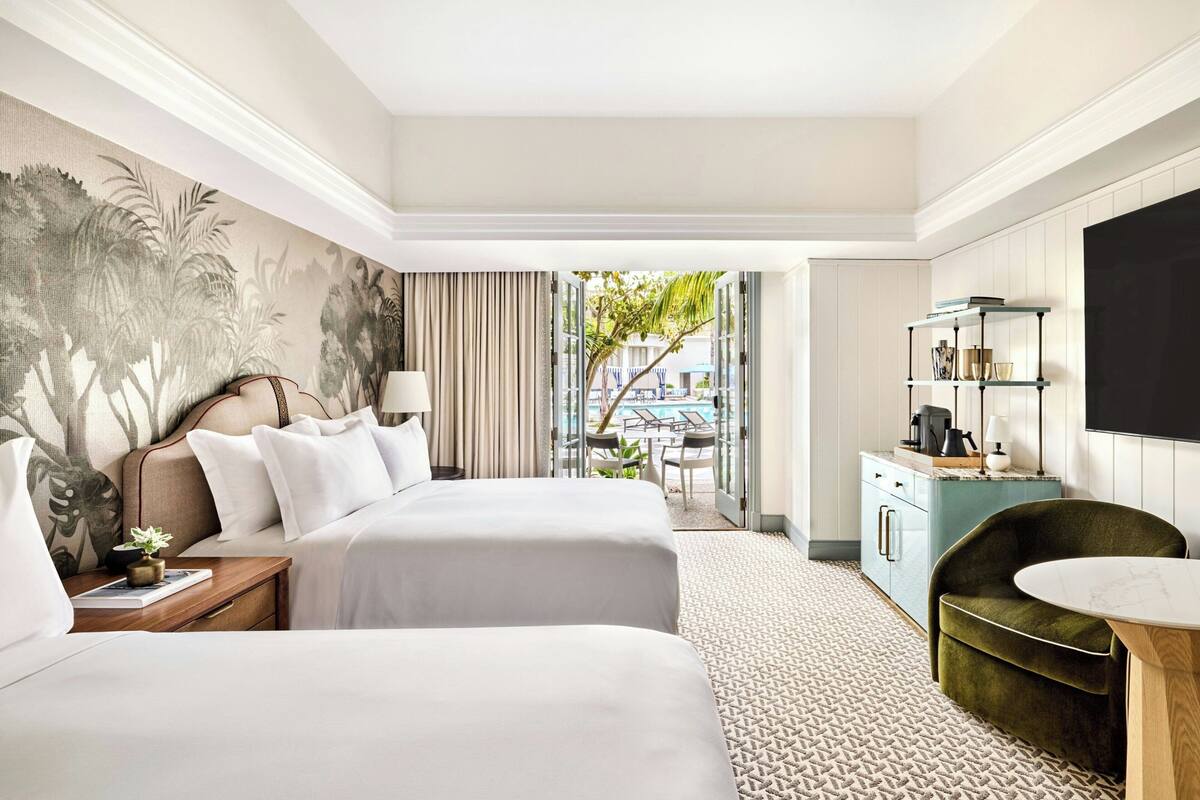 A hotel room with two beds, a desk, a chair, a TV, and a view of the pool.