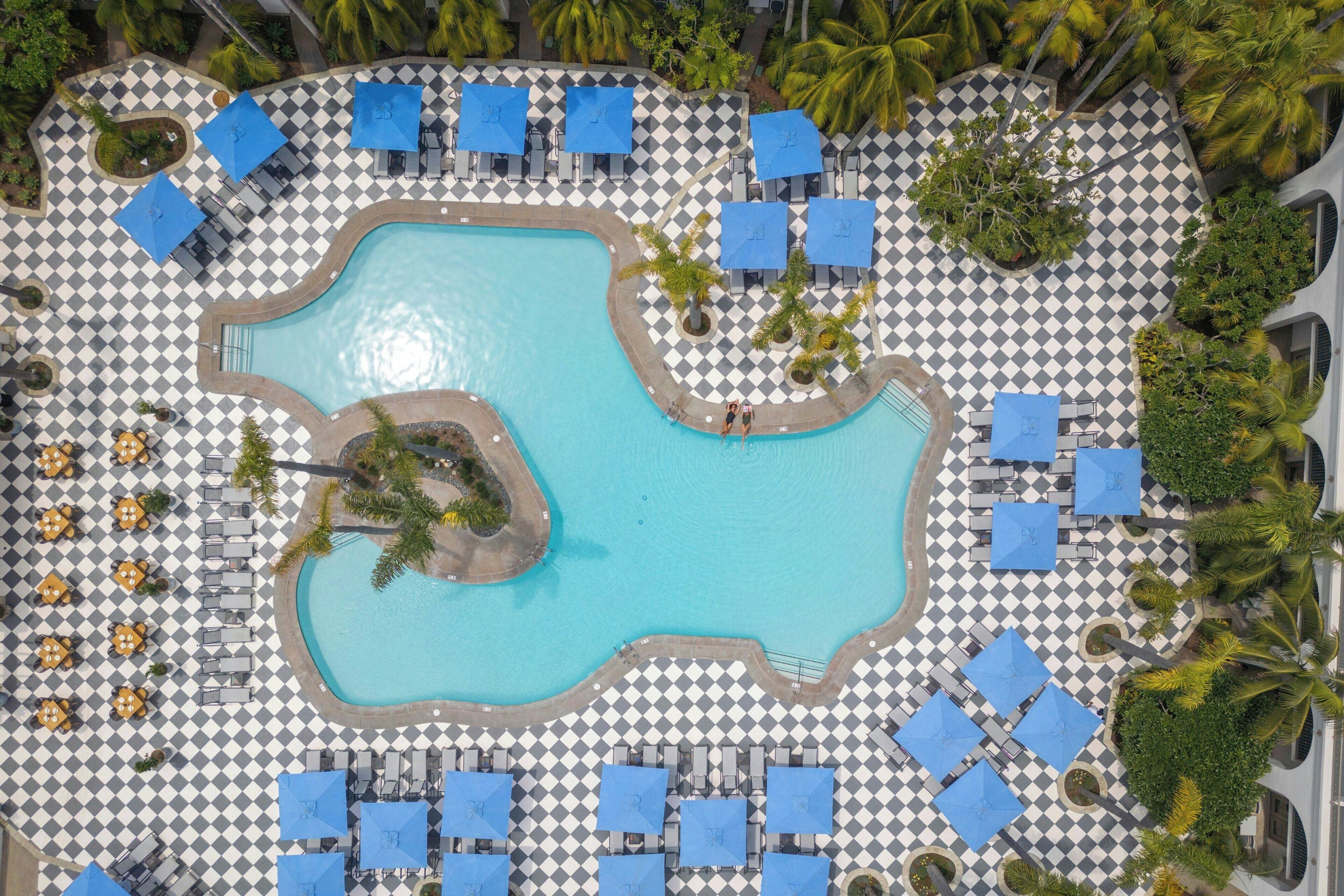 Vista aérea de un complejo turístico con una piscina central rodeada de suelo a cuadros, sillones de descanso y sombrillas.