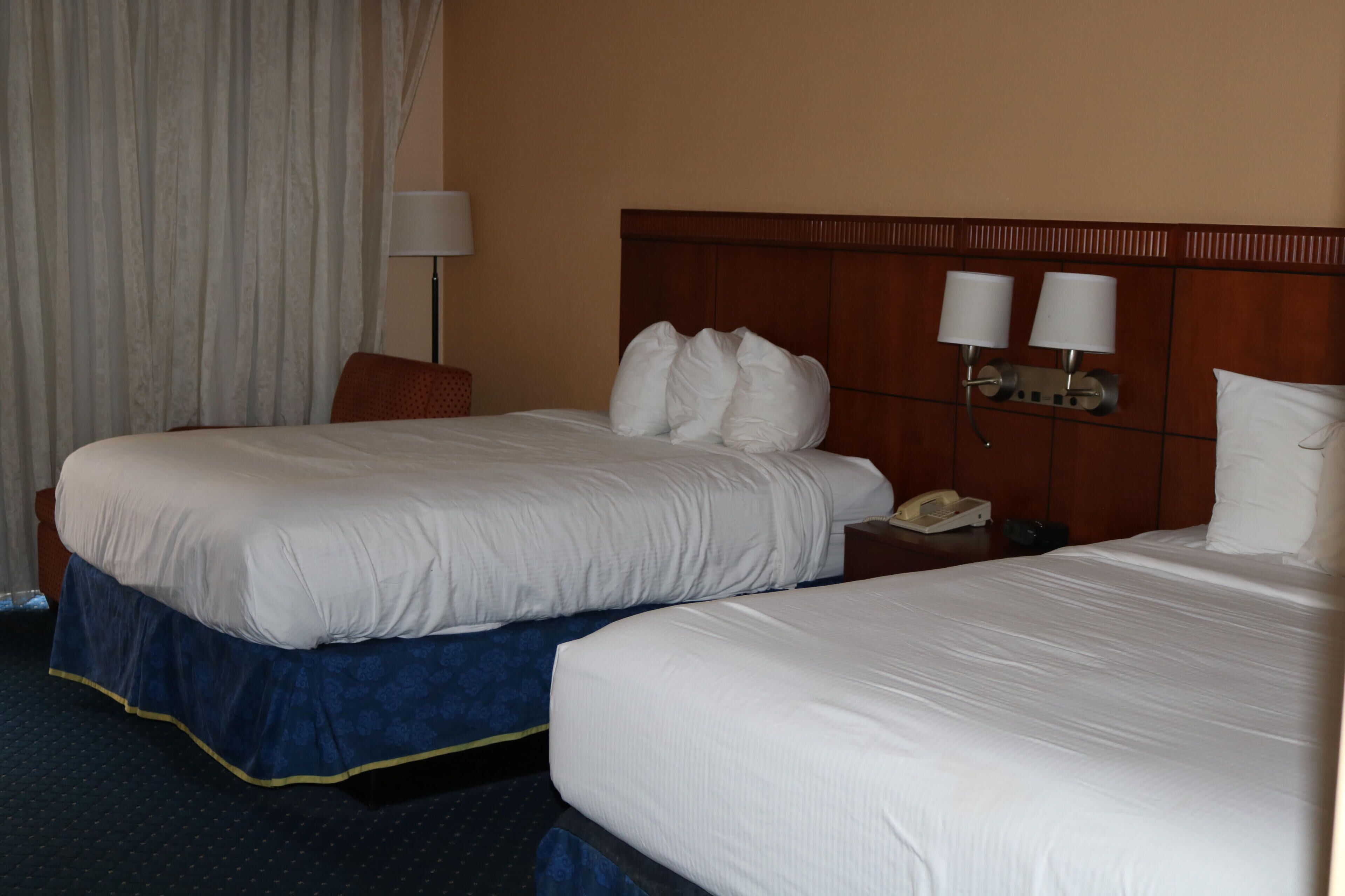 A hotel room with two beds, a nightstand with a phone, and a lamp.