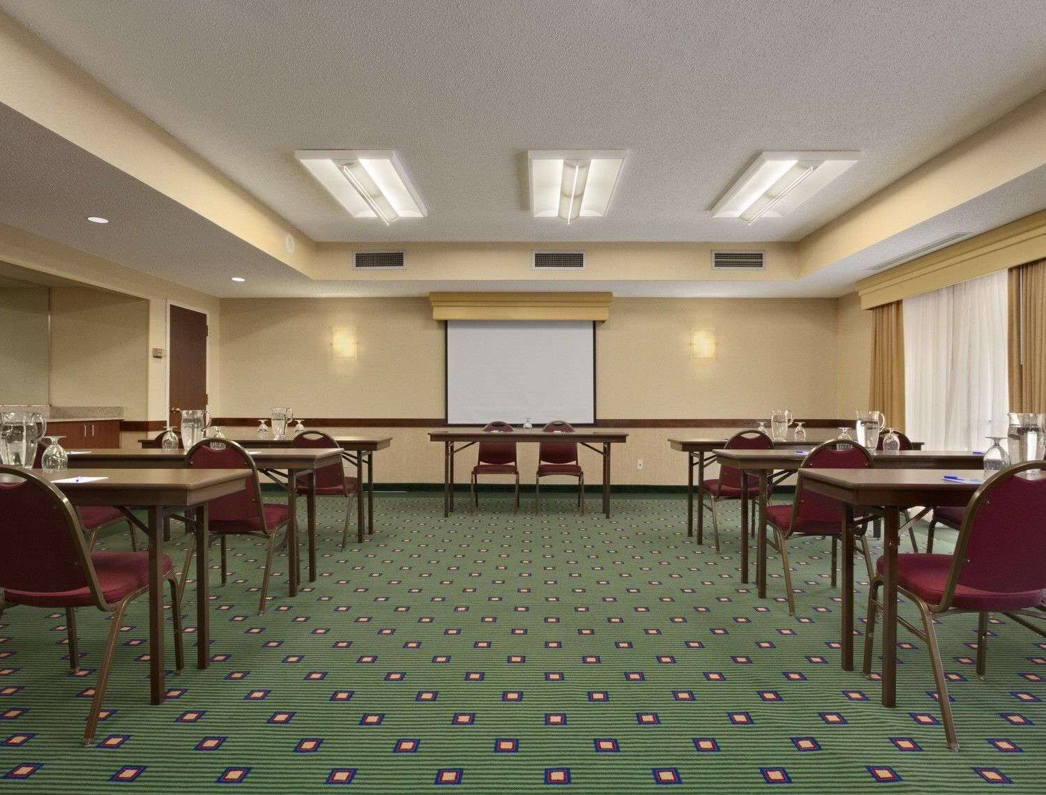 A conference room with a green patterned carpet, multiple tables with chairs, a projector screen, and ceiling lights.