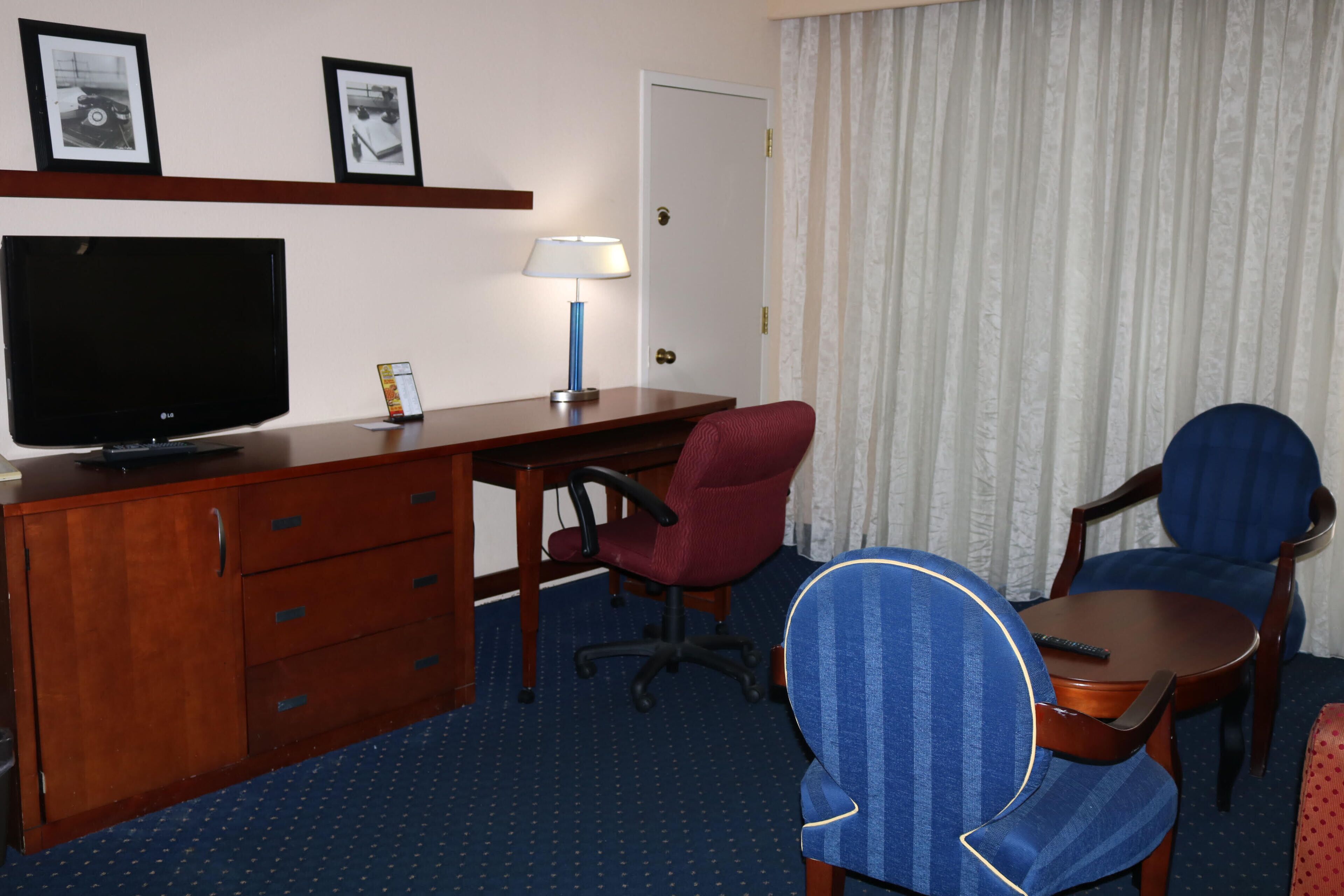A hotel room with a wooden desk, a TV, a lamp, two chairs, and a bed.