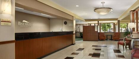 A hotel reception area with a Baymont Inn & Suites sign, a reception desk, a clock, and a seating area with chairs and a table.