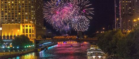 A nighttime cityscape with fireworks over a river, illuminated buildings, and a boat.