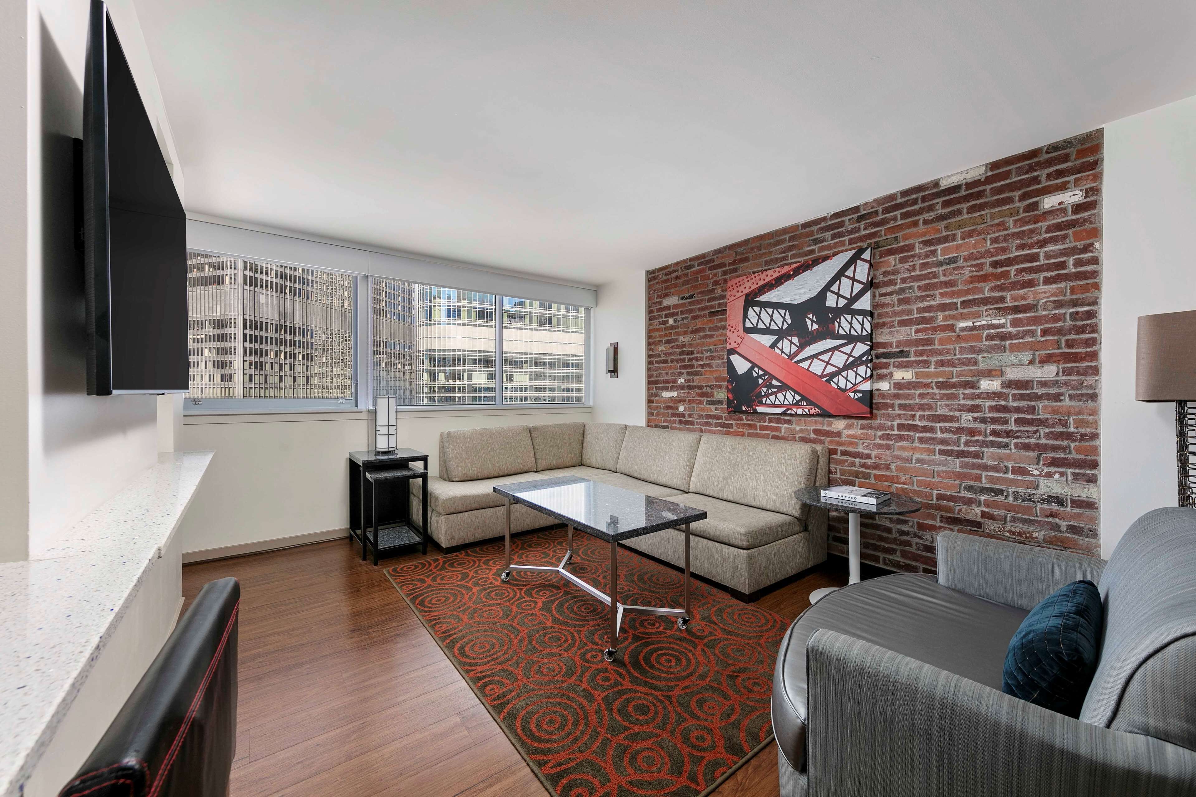 A modern living room with a sofa, a glass coffee table, a patterned rug, and a brick accent wall.
