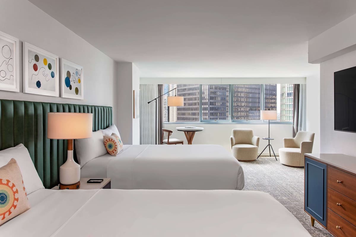 A hotel room with two beds, a green headboard, a desk, a chair, a lamp, and a view of a cityscape through large windows.