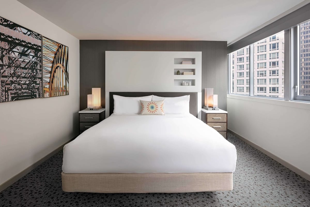 A hotel room with a large bed, two bedside tables, a wall-mounted shelf, and a window with a city view.