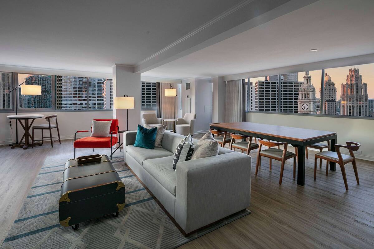 A modern hotel room with a city view, a sofa, a dining table, and a suitcase.