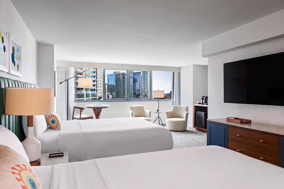 A hotel room with two beds, a TV, a desk, and a view of the cityscape.