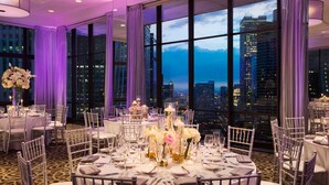 A high-rise hotel room with large windows offering a city view, round tables set for dining, and elegant decor with flowers and candles. - The Royal Sonesta Chicago Downtown (Chicago)