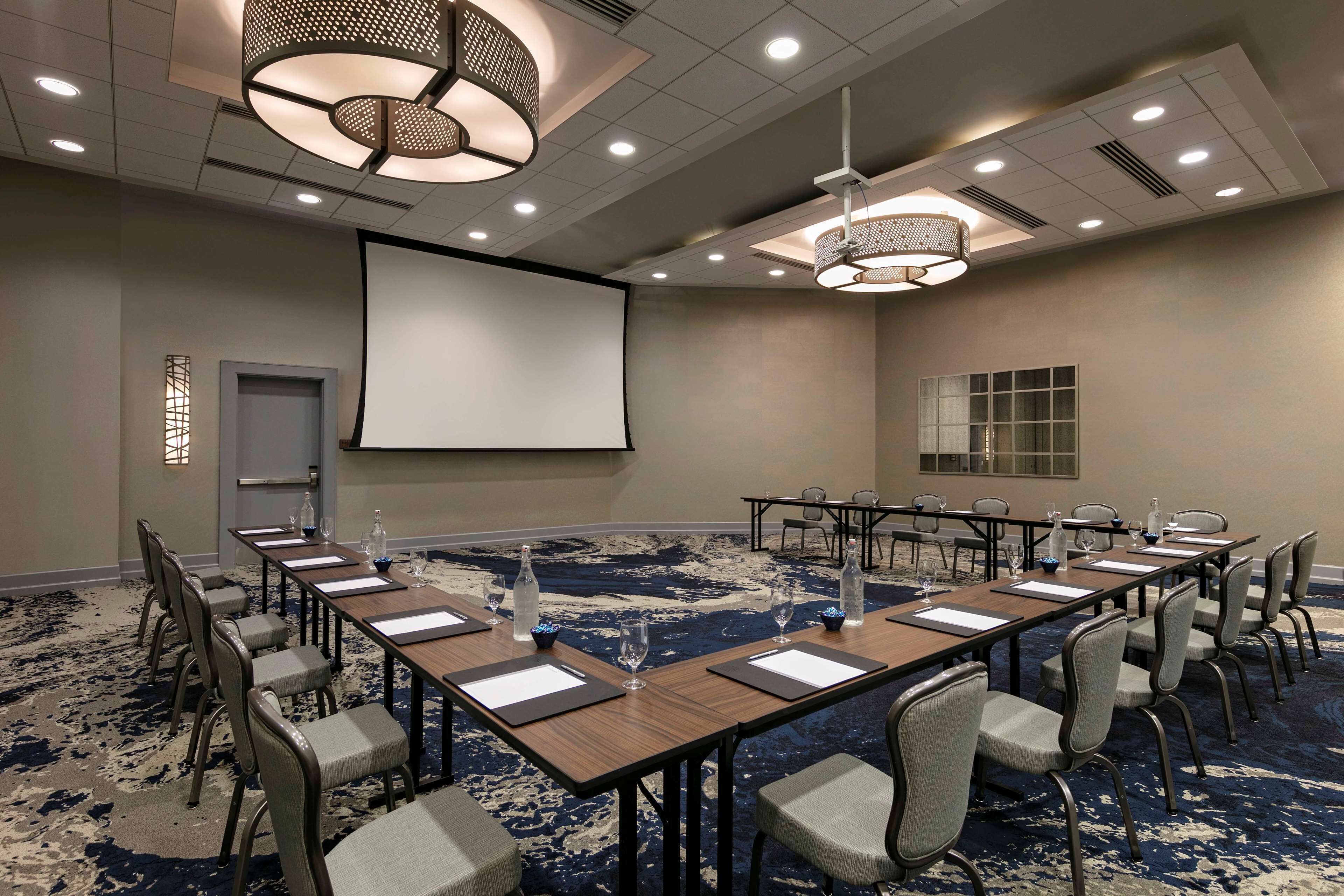 A conference room with a large screen, long tables, chairs, and a carpeted floor.