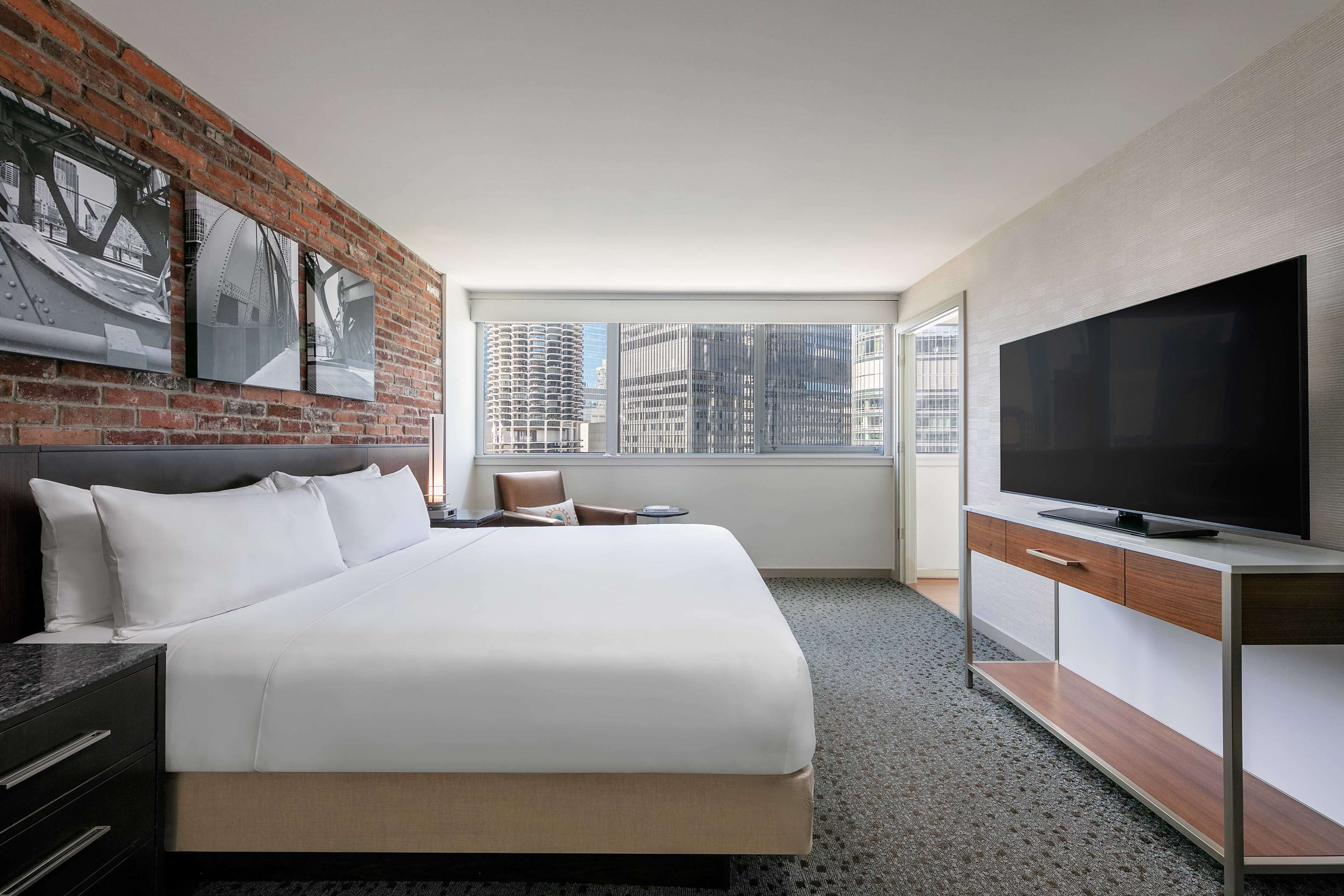 Una habitación de hotel moderna con una cama grande, un televisor, un escritorio y una pared de ladrillo decorada con obras de arte.