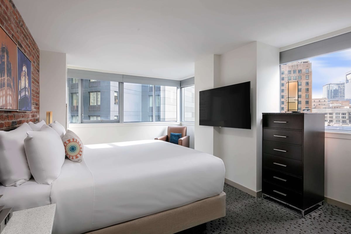 A hotel room with a large bed, a TV mounted on the wall, a dresser, and a window with a city view.