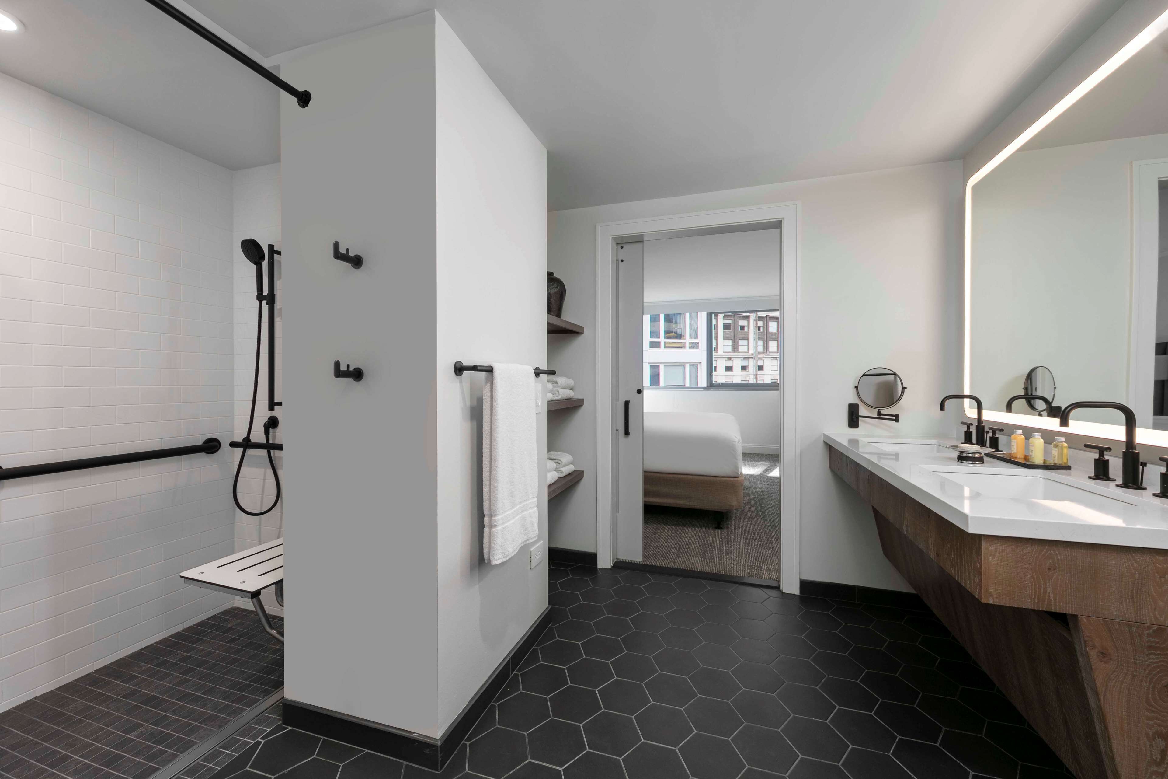 Ein modernes Badezimmer mit Dusche, Waschtisch und Spiegel, mit Blick auf ein Schlafzimmer durch eine offene Tür.