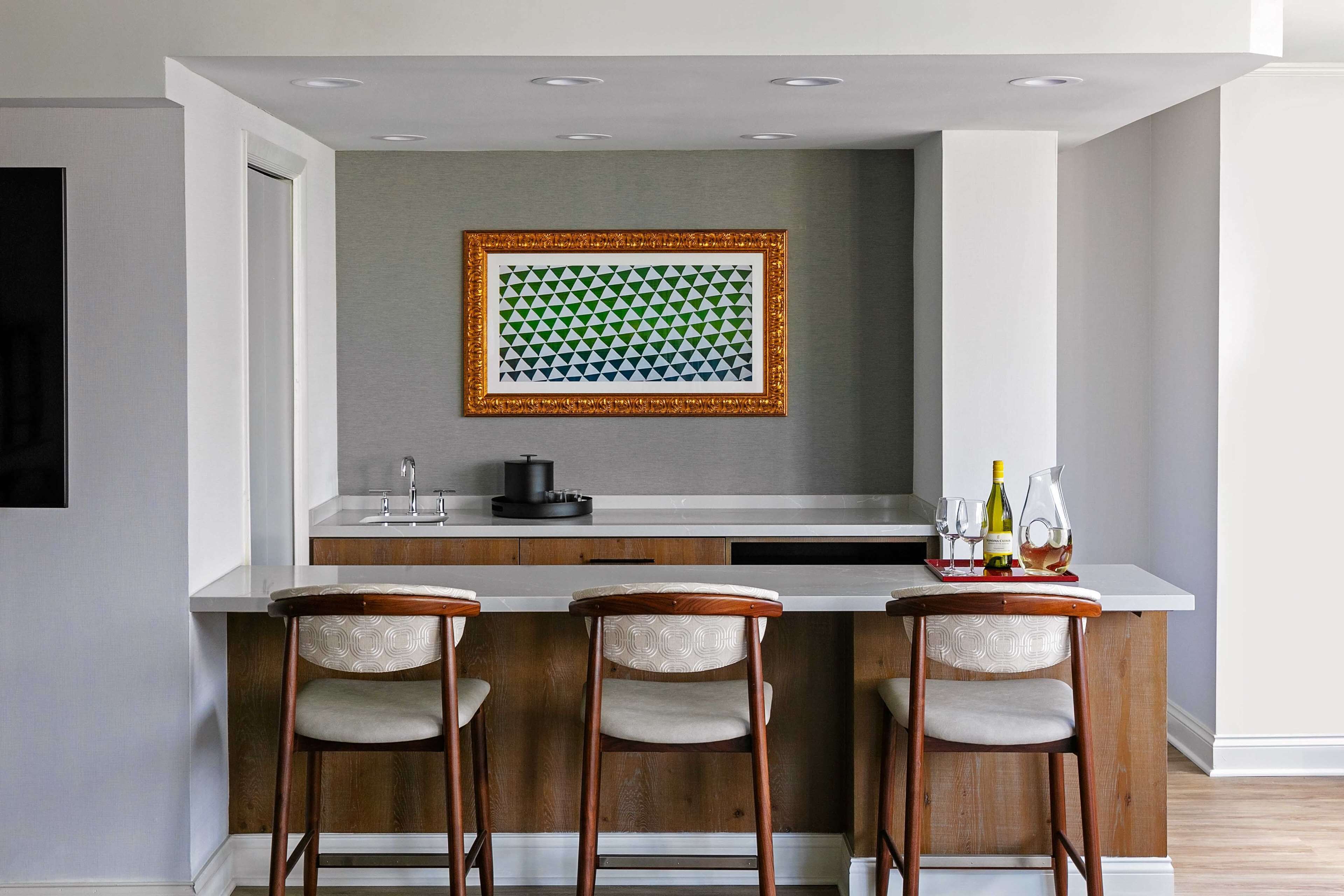 A modern kitchen with a white countertop, three wooden bar stools, and a framed geometric pattern on the wall.