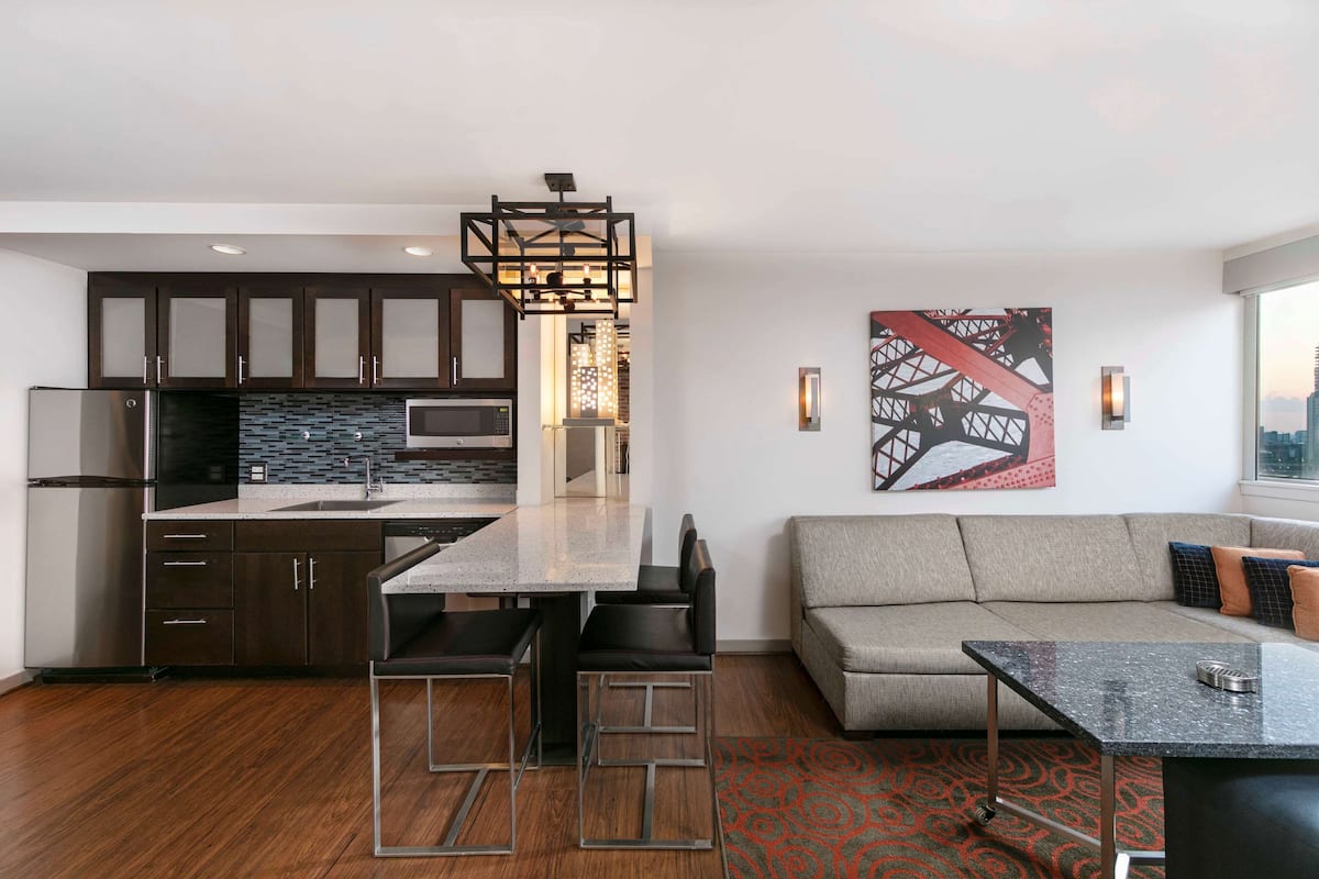 A modern kitchen with dark cabinets, a stainless steel refrigerator, and a microwave. There's a dining area with a table and chairs, and a living room with a sofa and a patterned rug.