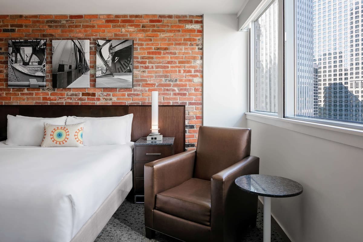 A hotel room with a brick wall, a bed with white bedding, a brown leather chair, a small round table, and a nightstand with a lamp.
