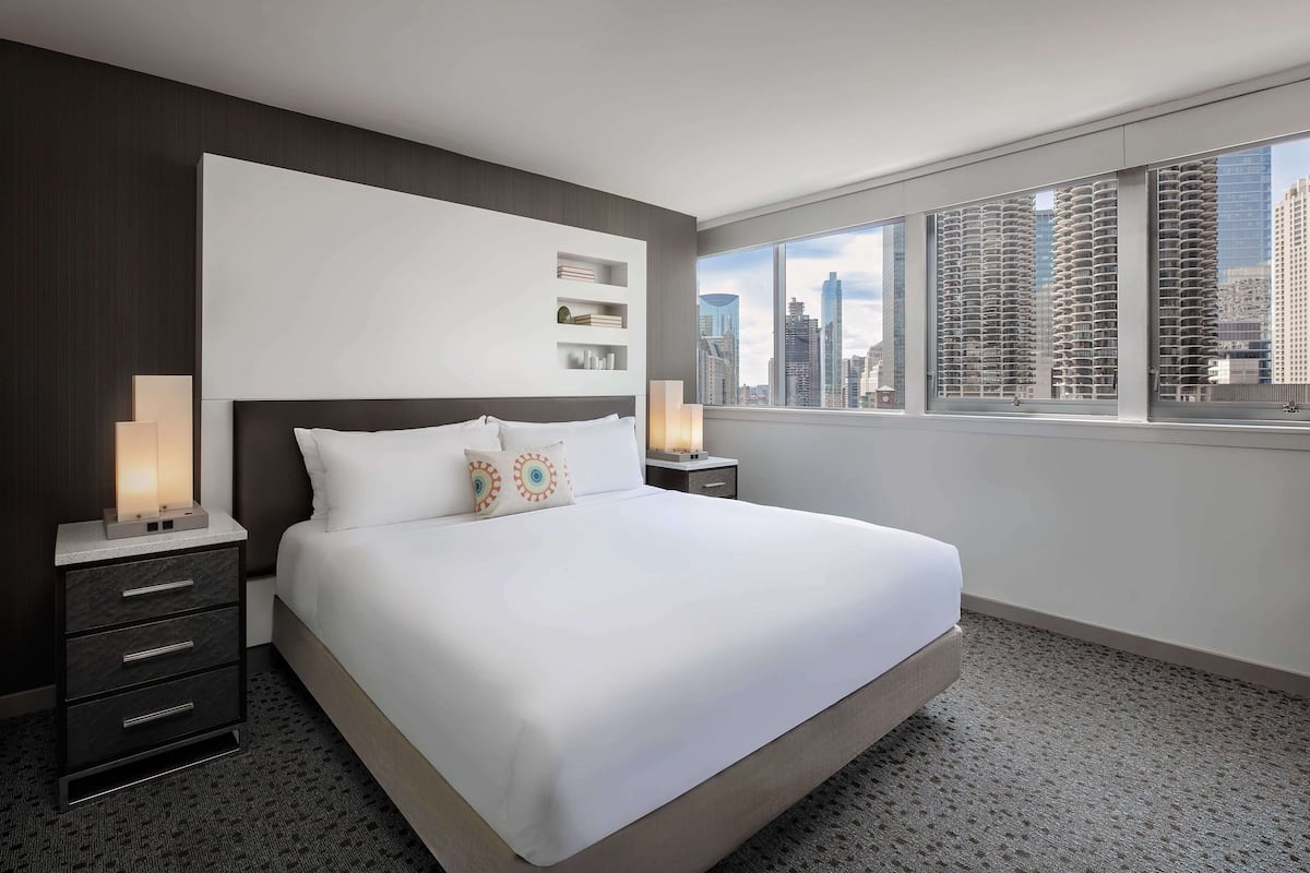A modern hotel room with a large bed, dark wood paneling, a city view, and a nightstand with a lamp.