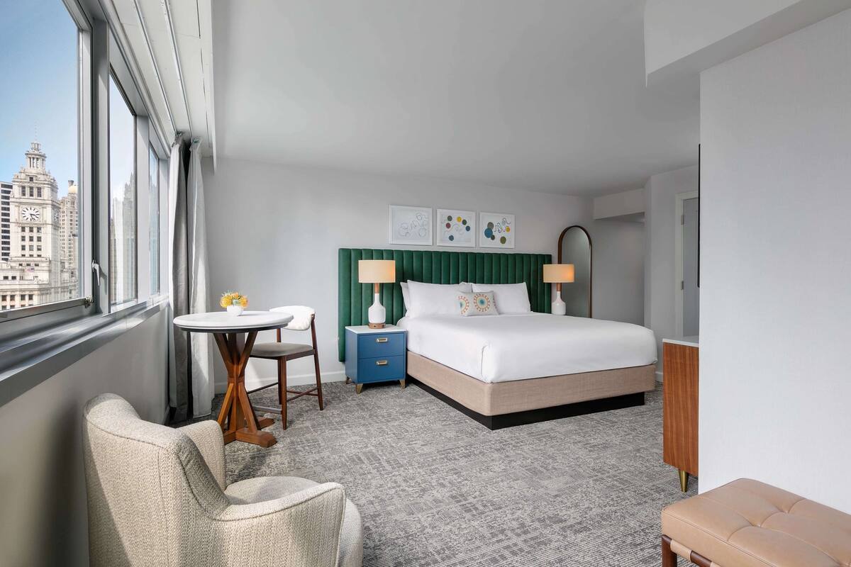 A hotel room with a large bed, a desk, a chair, and a view of a cityscape.
