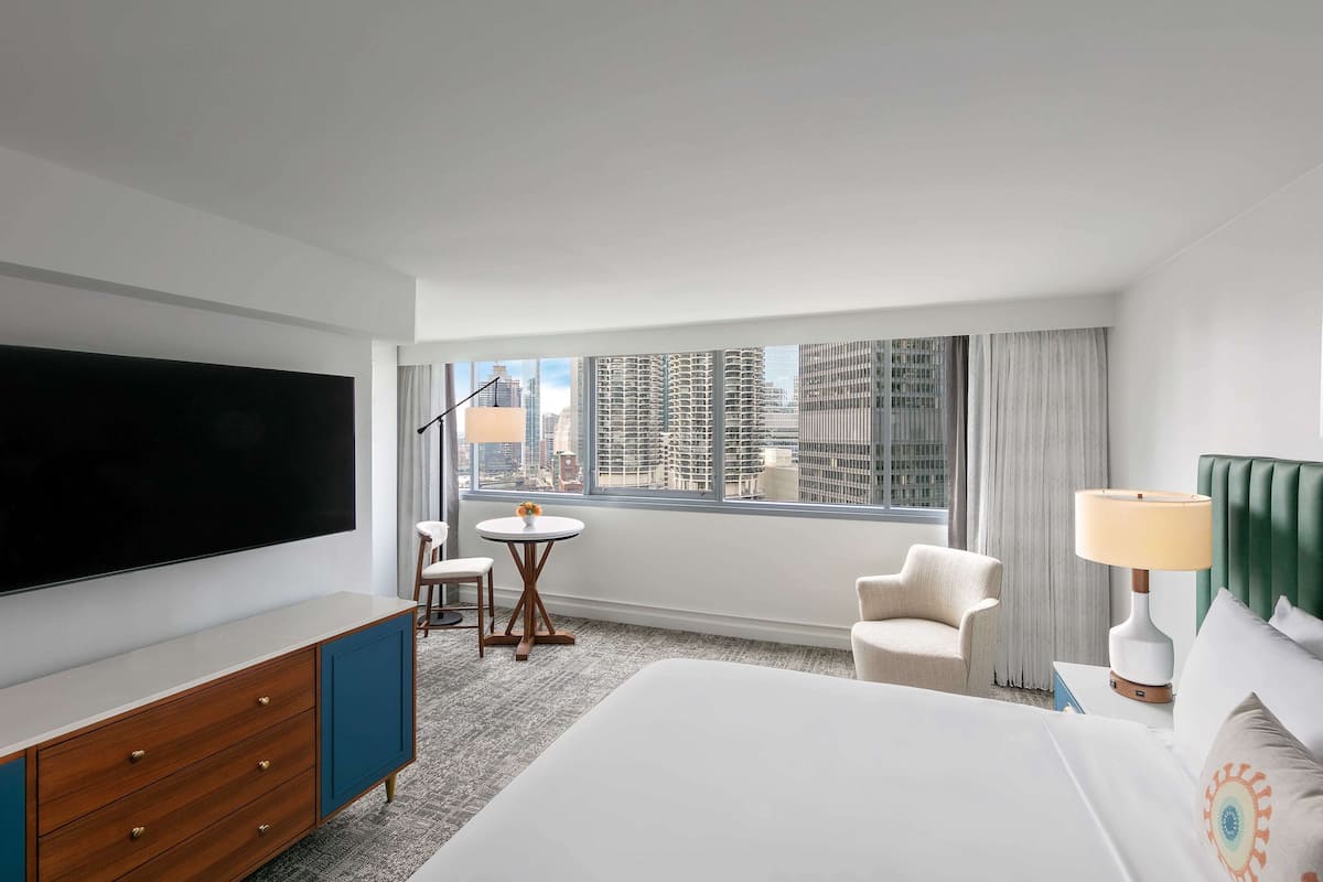 A hotel room with a large bed, a flat-screen TV, a desk with a lamp, a chair, and a view of the city through large windows.