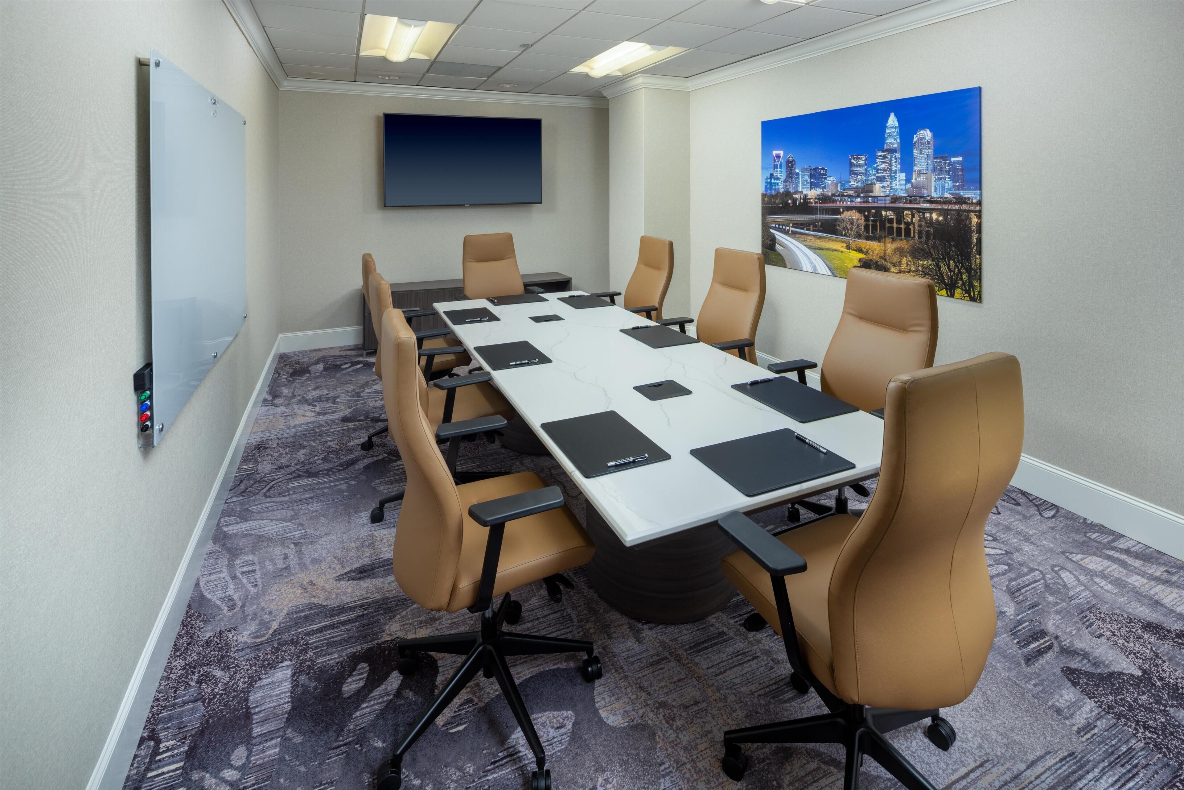 Un salón de conferencias con una larga mesa, sillas color beige, una pantalla de proyector y un cuadro de un paisaje urbano.