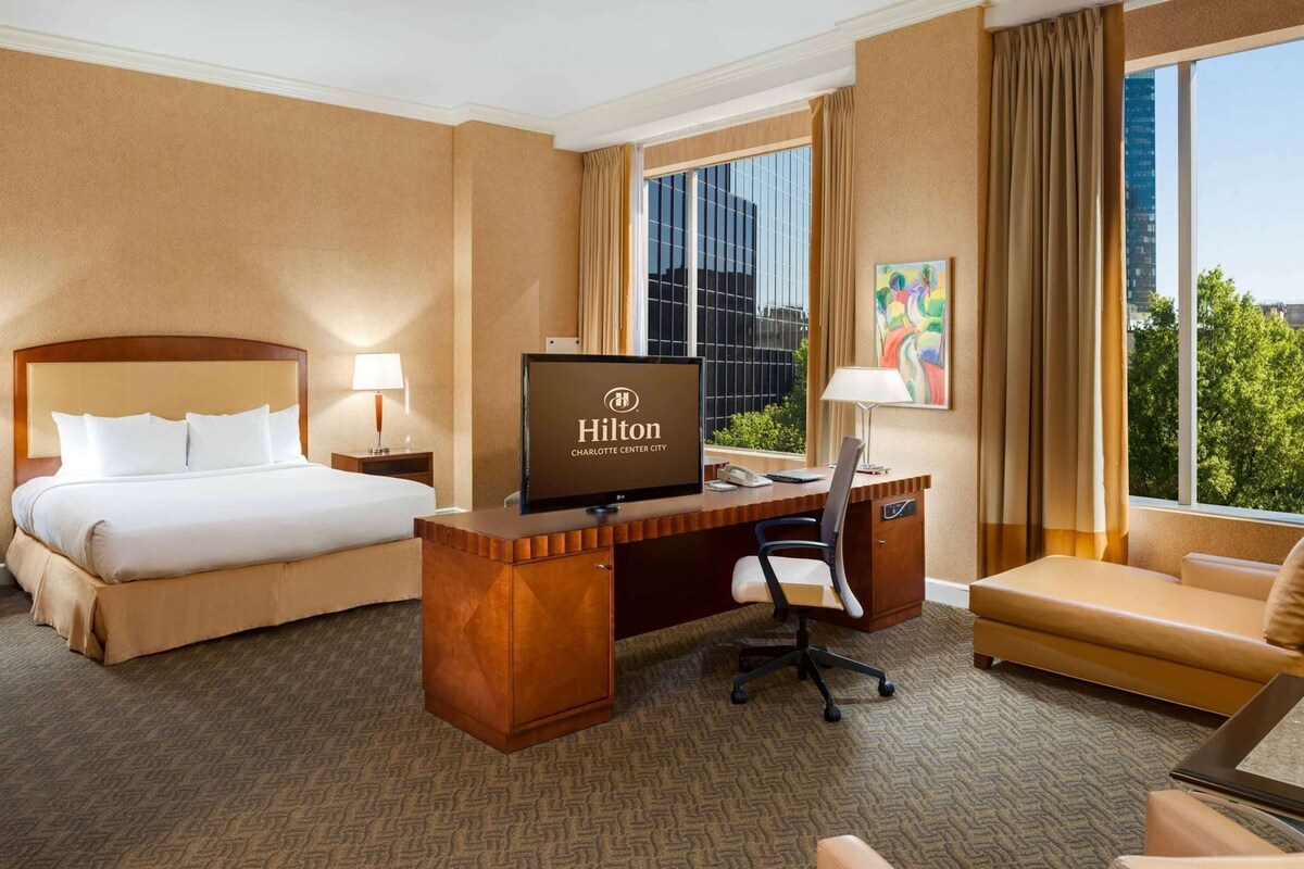 A hotel room with a bed, desk, chair, and a TV displaying the Hilton Charlotte Center City logo.