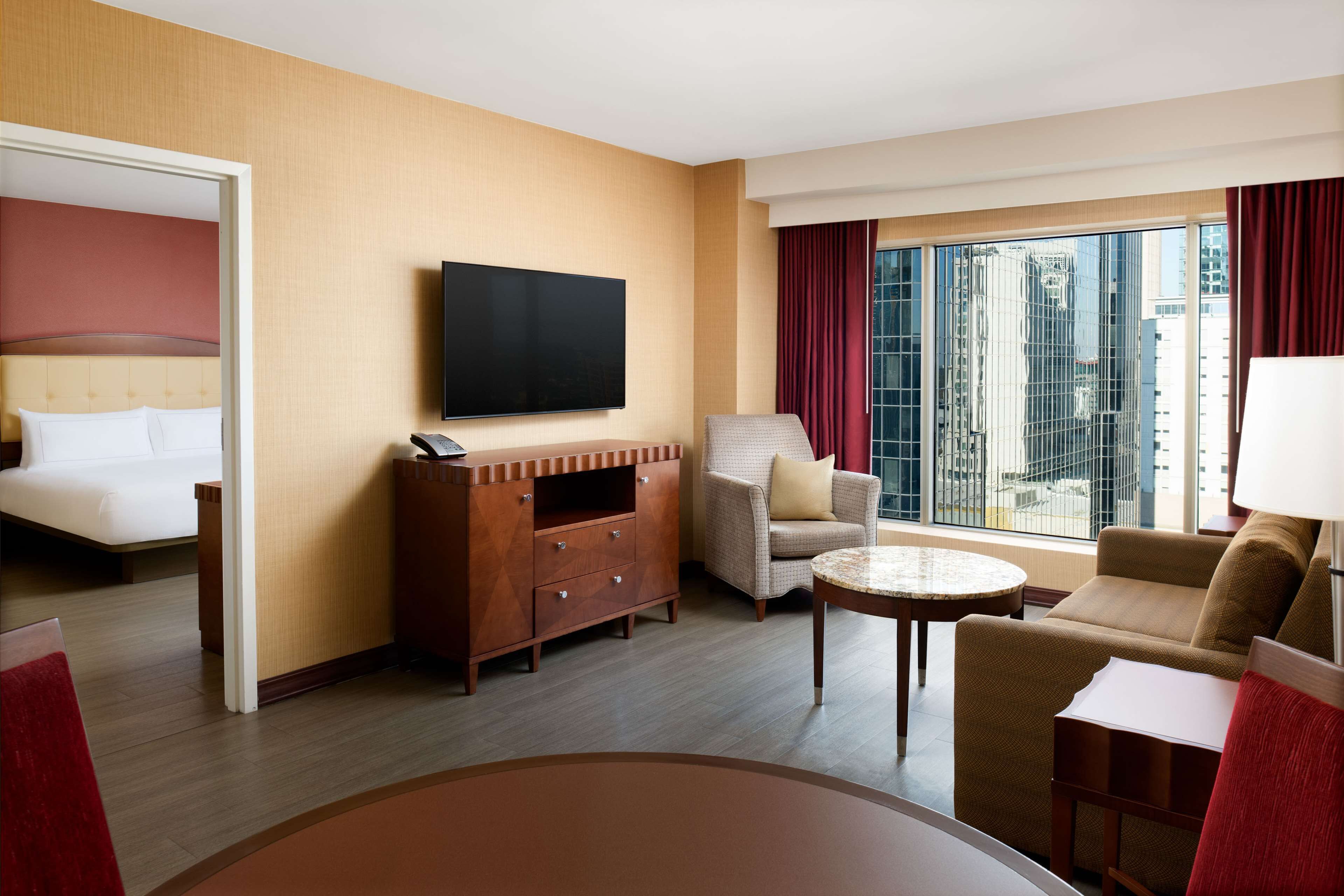A hotel room with a bed, a TV, a sofa, a chair, a small table, and a large window with a city view.