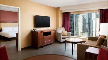 A hotel room with a bed, a TV, a sofa, a chair, a small table, and a large window with a city view.