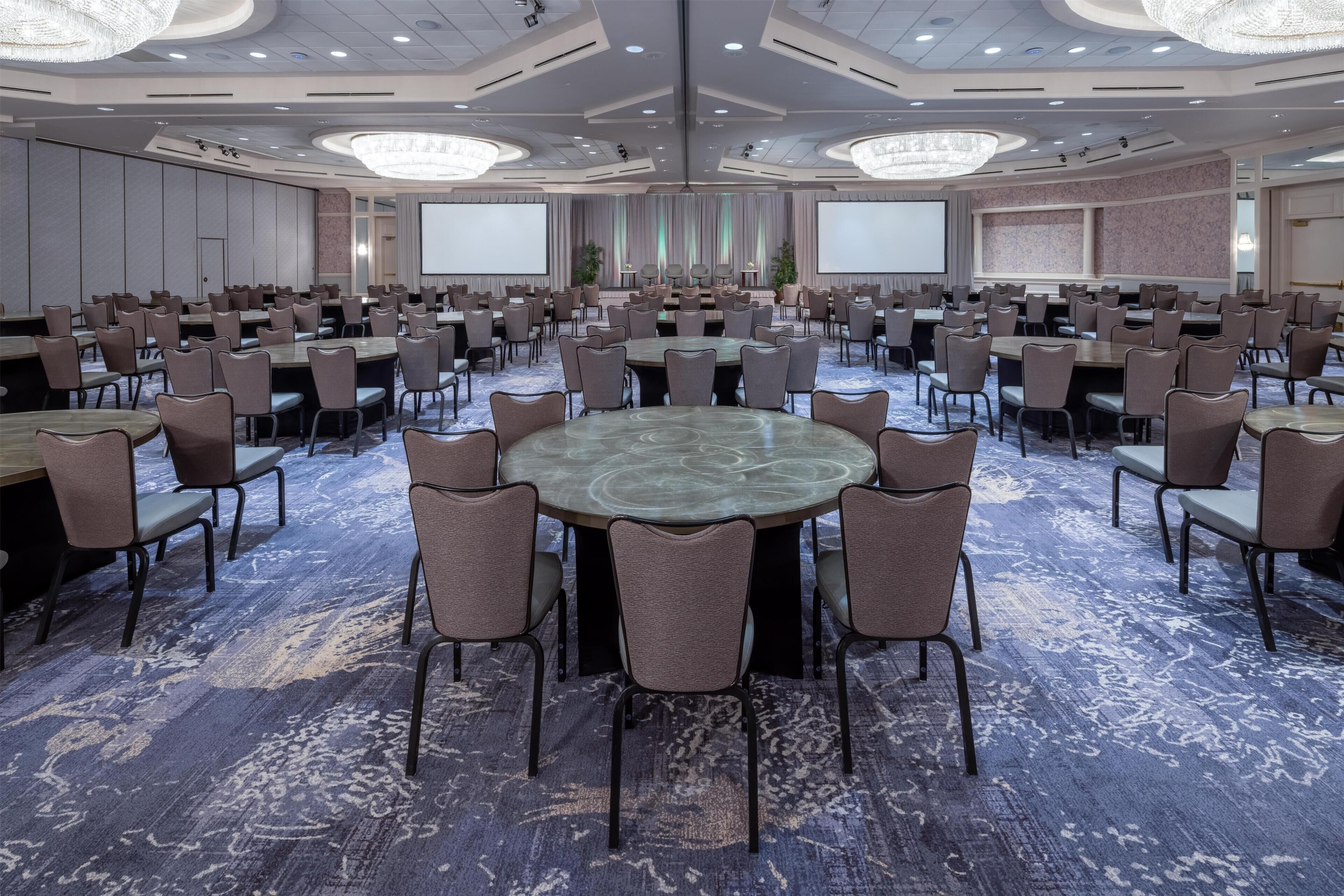 A conference room with multiple tables and chairs, large screens, and a carpeted floor.