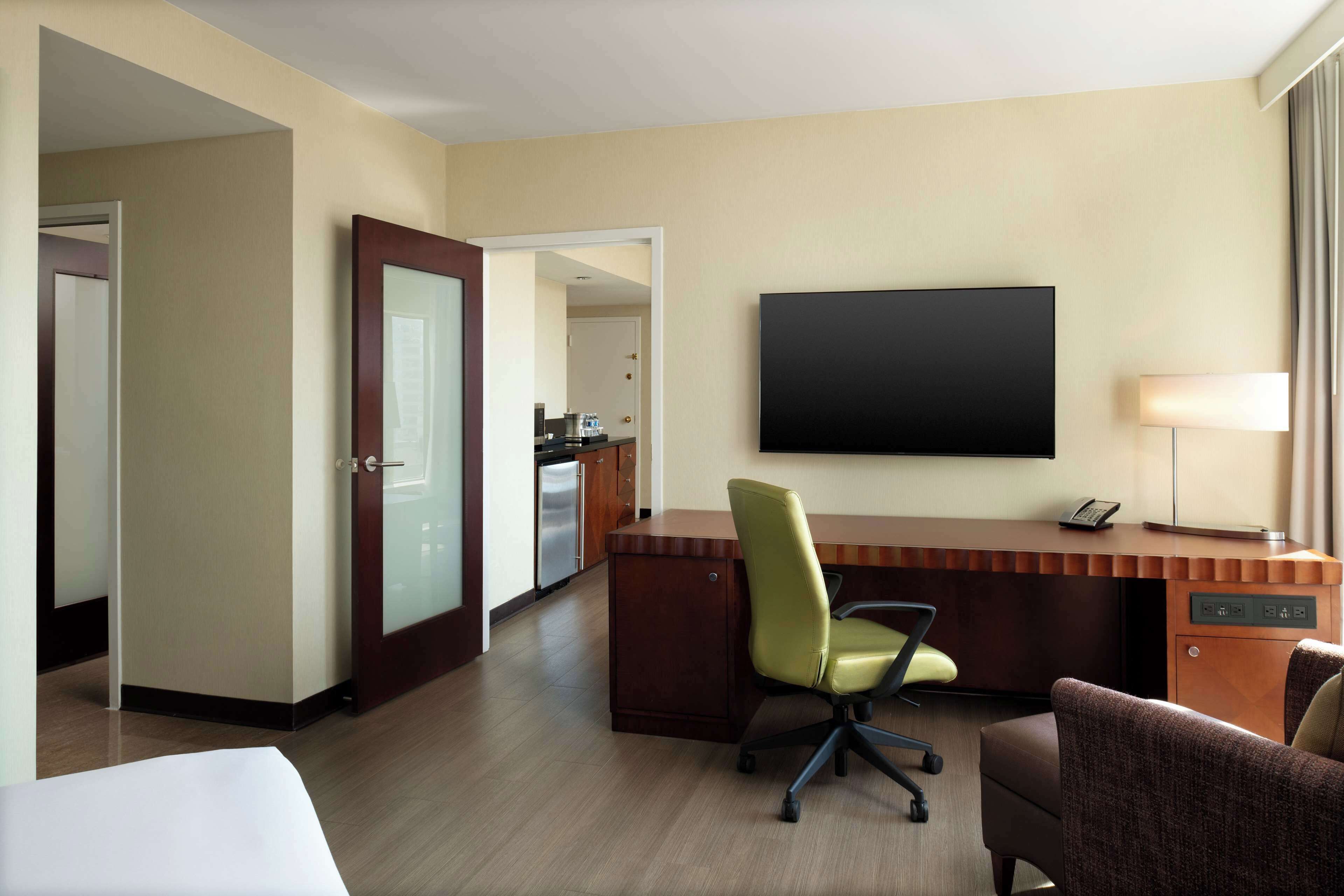 A hotel room with a wooden desk, a green office chair, a flat-screen TV, a lamp, and a bed.