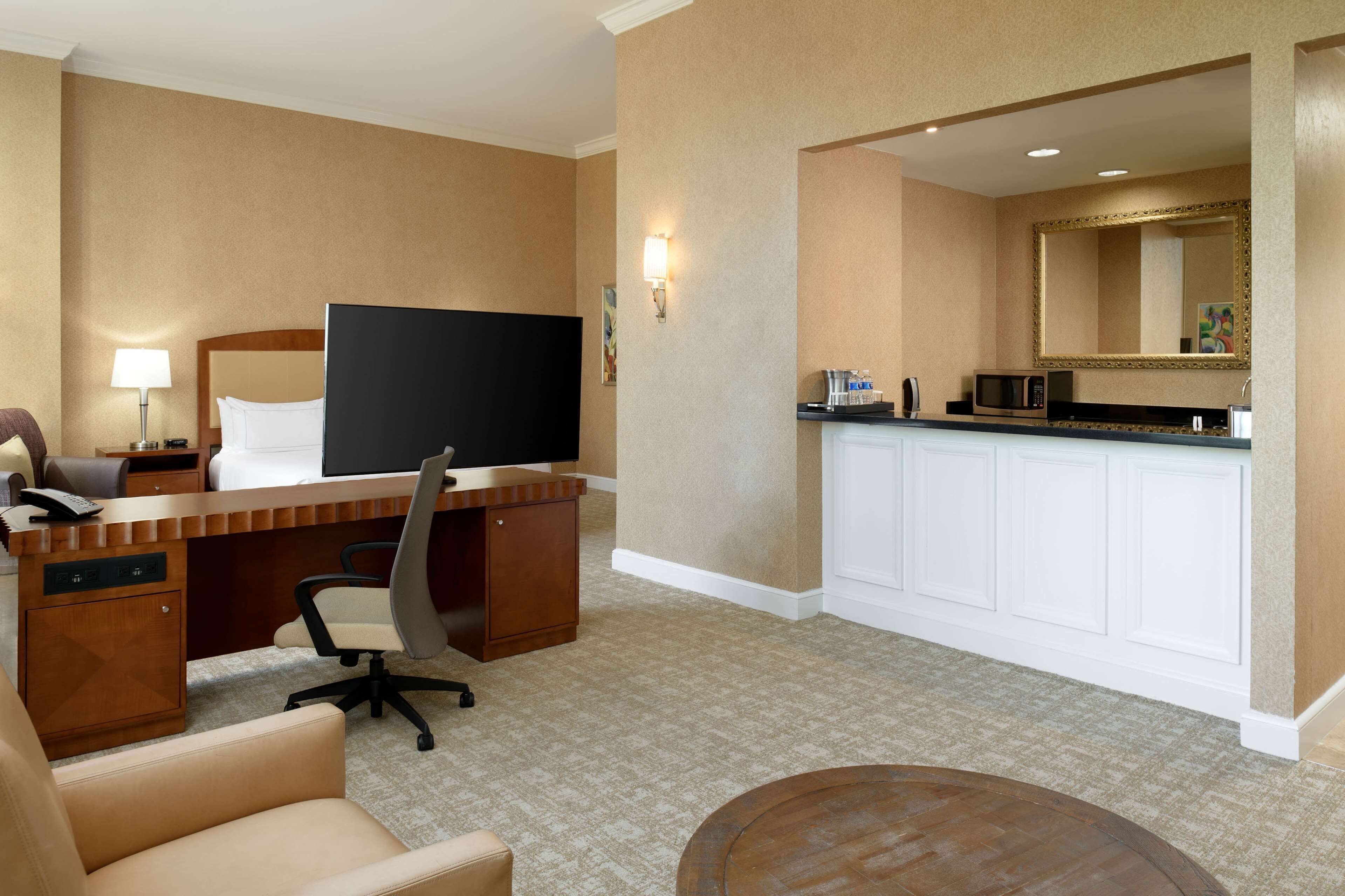 A hotel room with a bed, desk, chair, television, and a kitchenette area.