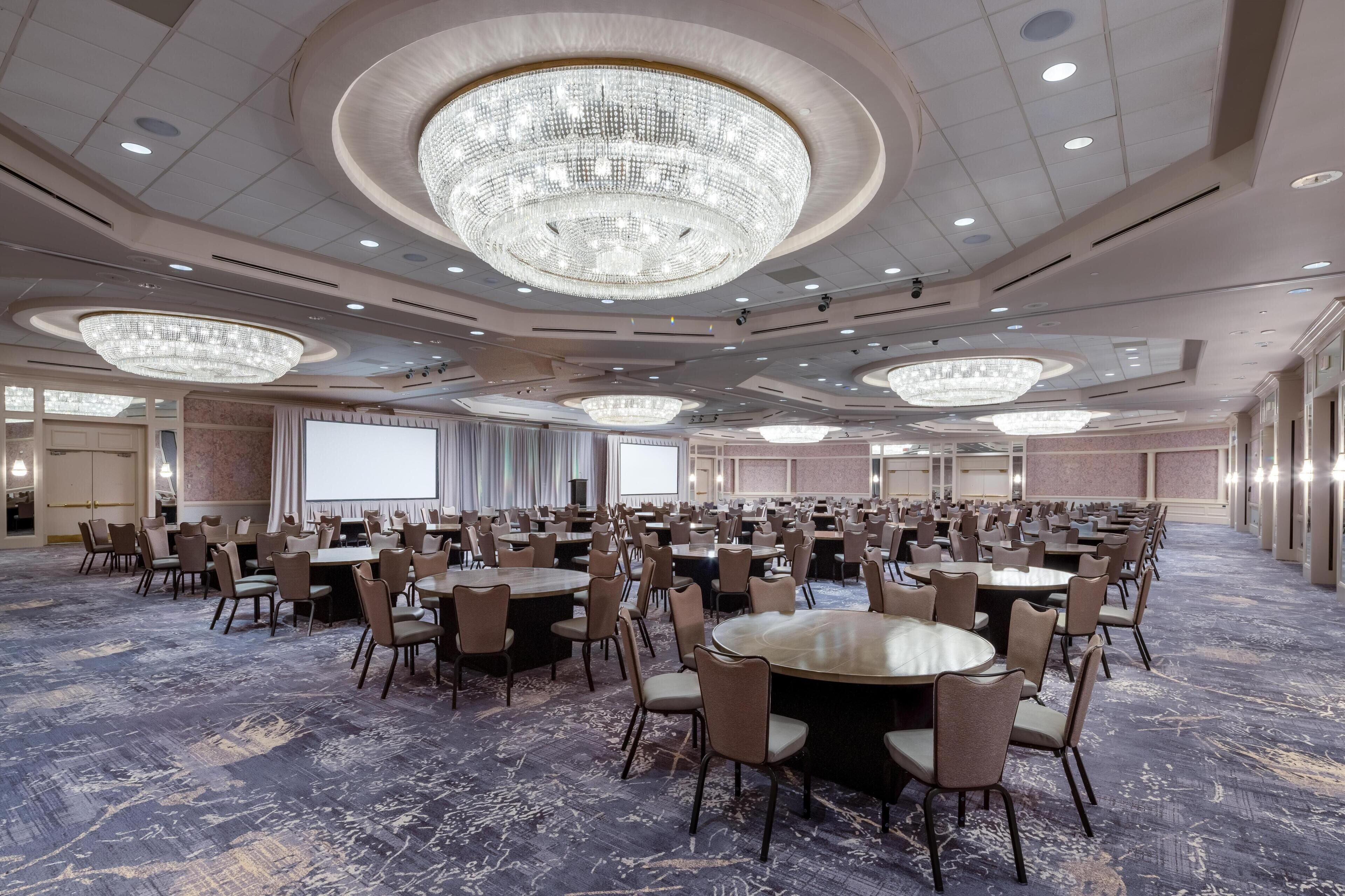 Une grande salle de conférence équipée de plusieurs tables et chaises, de lustres et d’un écran de projection.