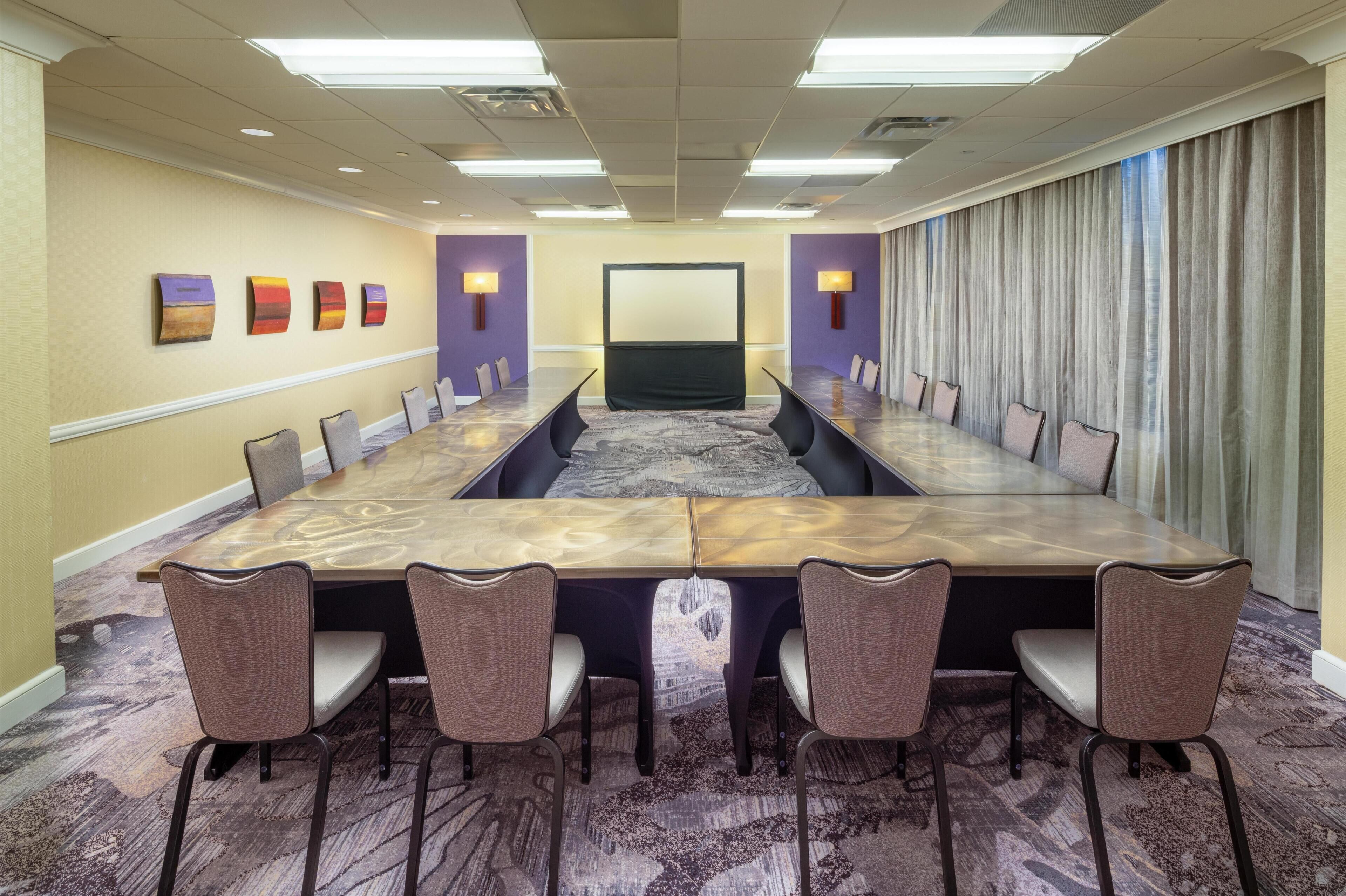 A conference room with a long table, chairs, a projector screen, and a large window with curtains.