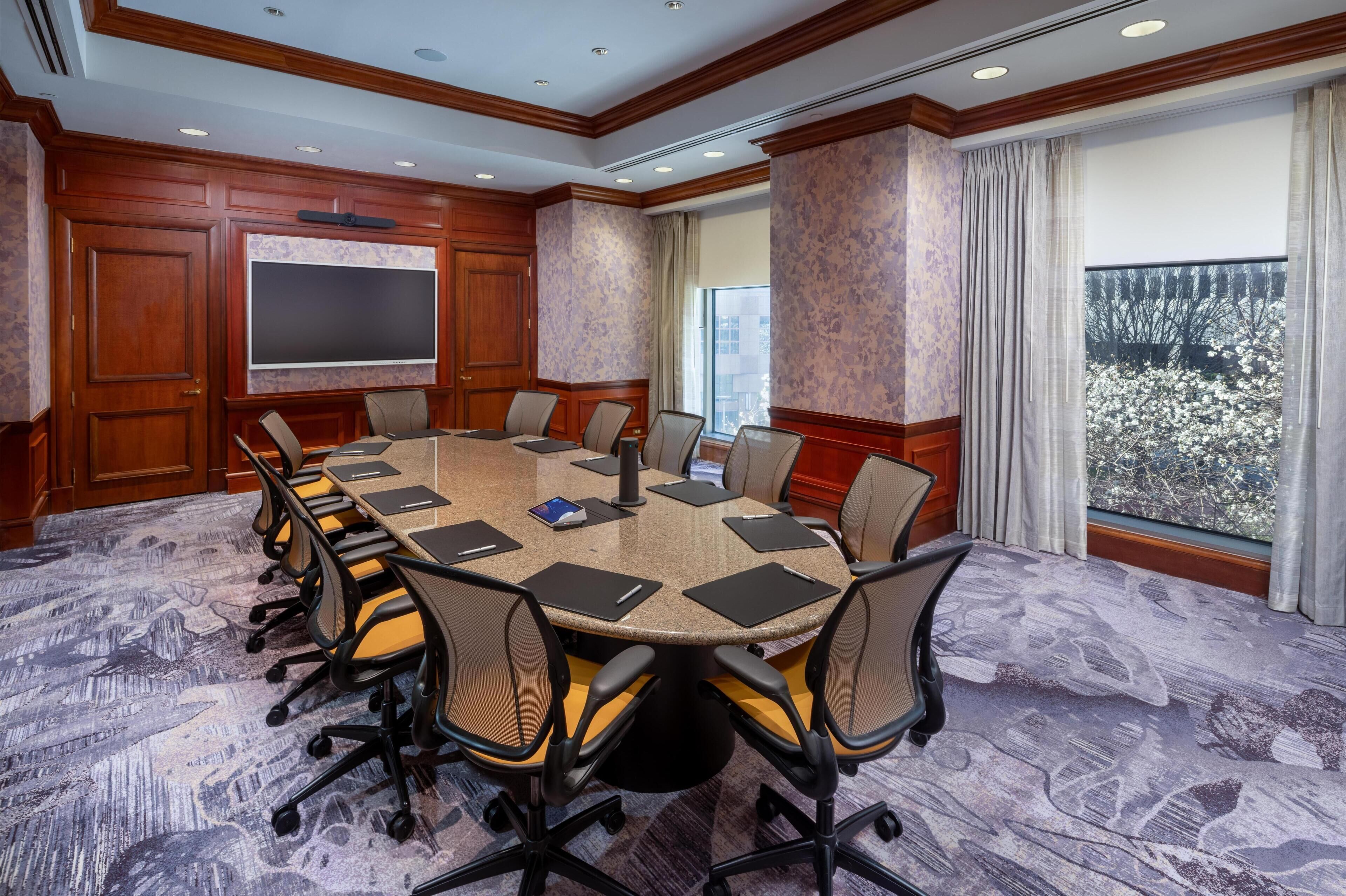 A conference room with a large table, chairs, a flat-screen TV, and a view of trees through the window.