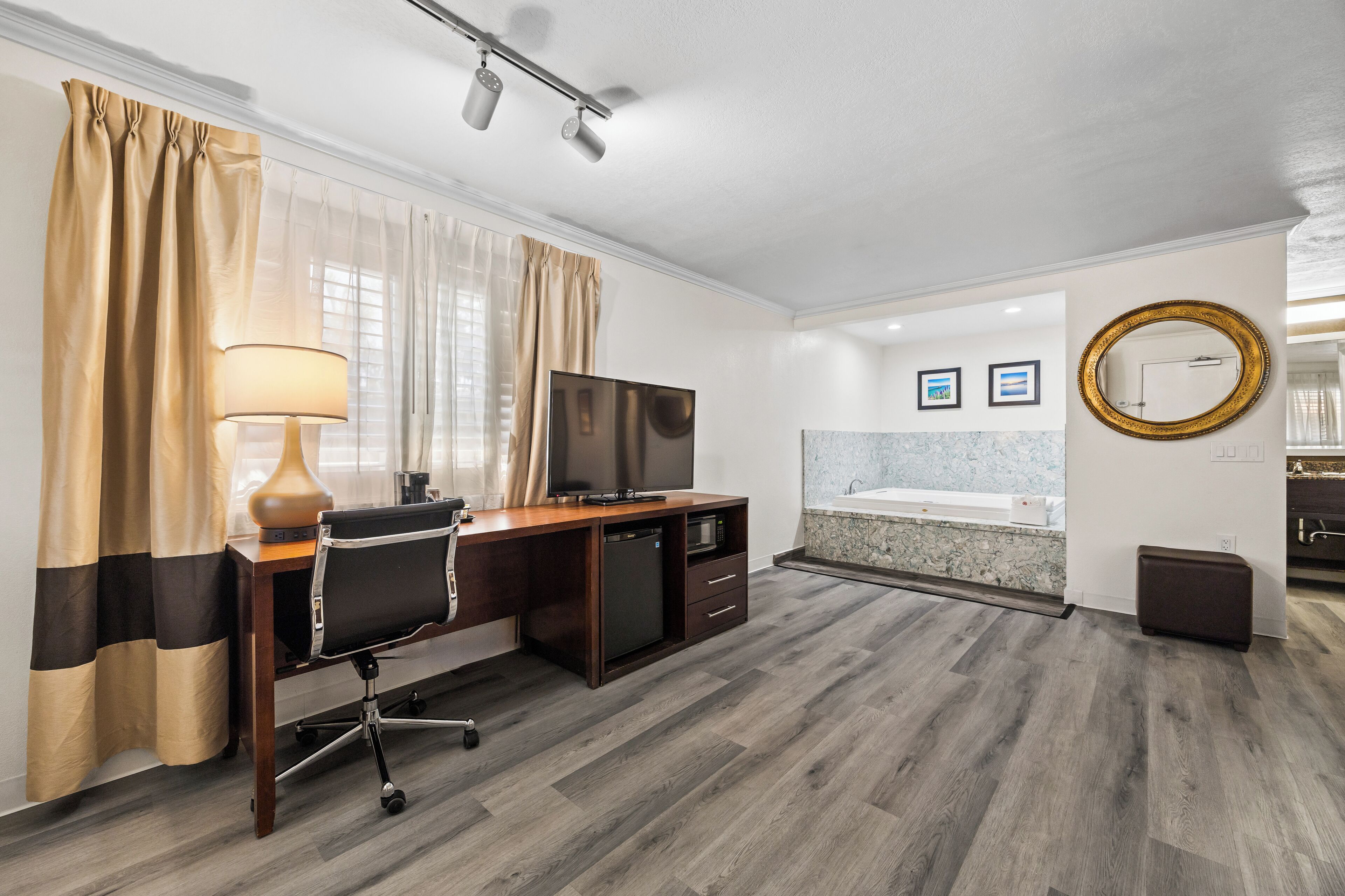 A hotel room with a wooden desk, a television, a lamp, a sofa, a small cabinet, a trash bin, and a bathroom with a bathtub and a marble countertop.