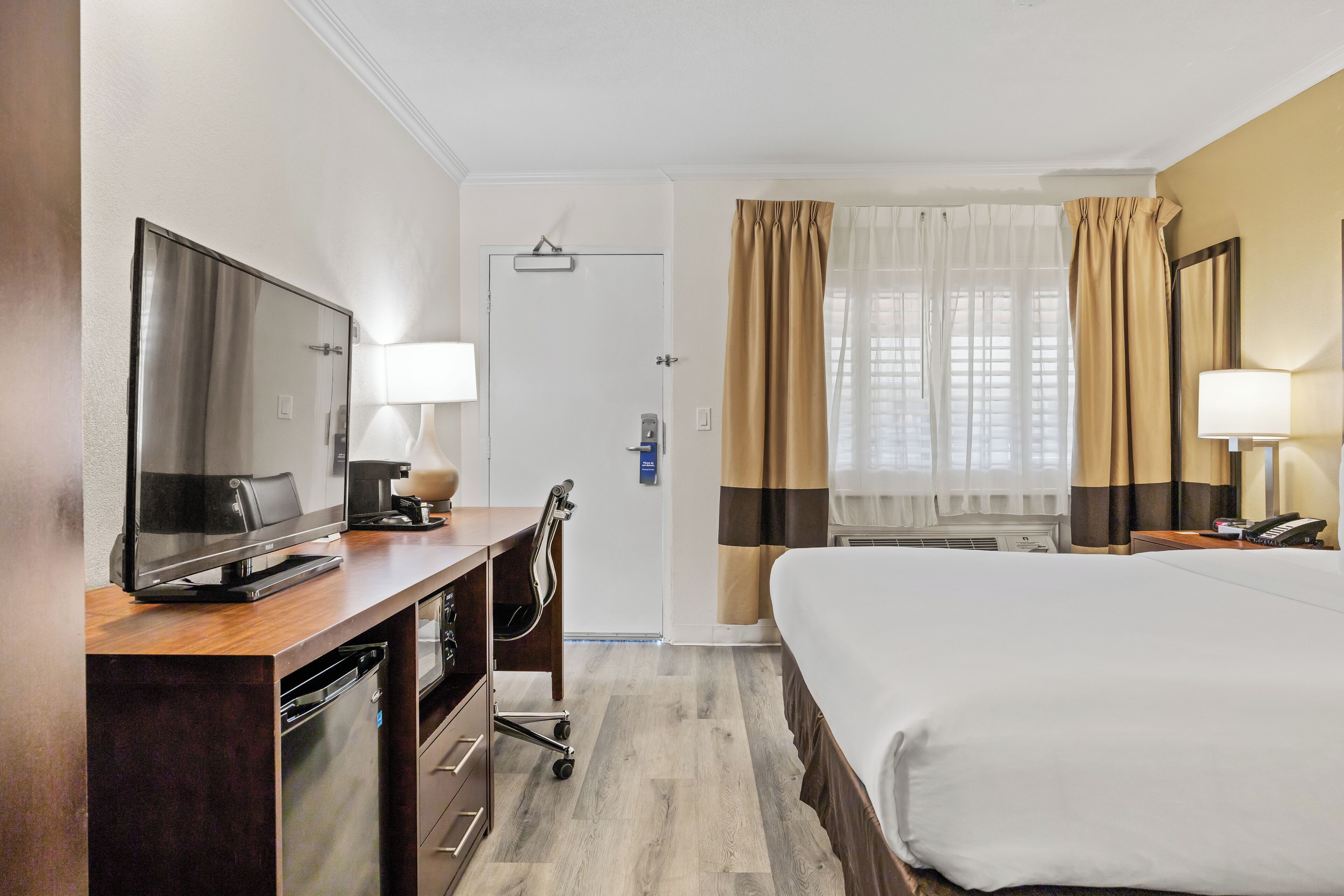 Habitación de hotel con cama, escritorio con televisor, lámpara de escritorio, silla, minibar y ventana con cortinas.