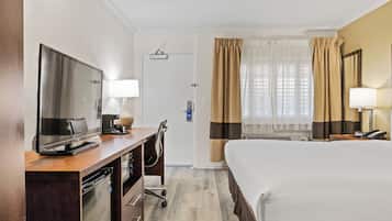 Quarto de hotel com uma cama, uma mesa de trabalho com televisão, luminária, cadeira, minibar e janela com cortinas.