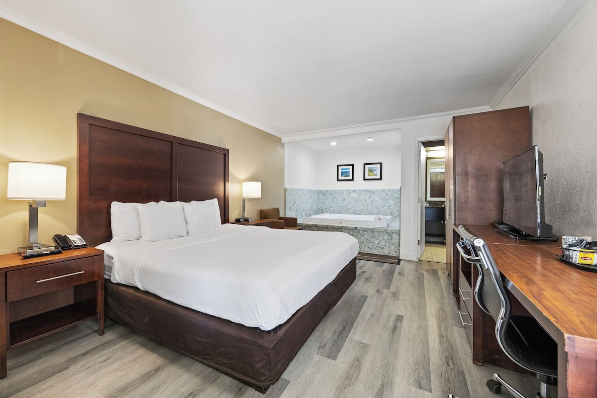 Ein Hotelzimmer mit einem großen Bett, Nachttischlampen, einem Schreibtisch mit Fernseher und einem Badezimmer mit Badewanne und Dusche.