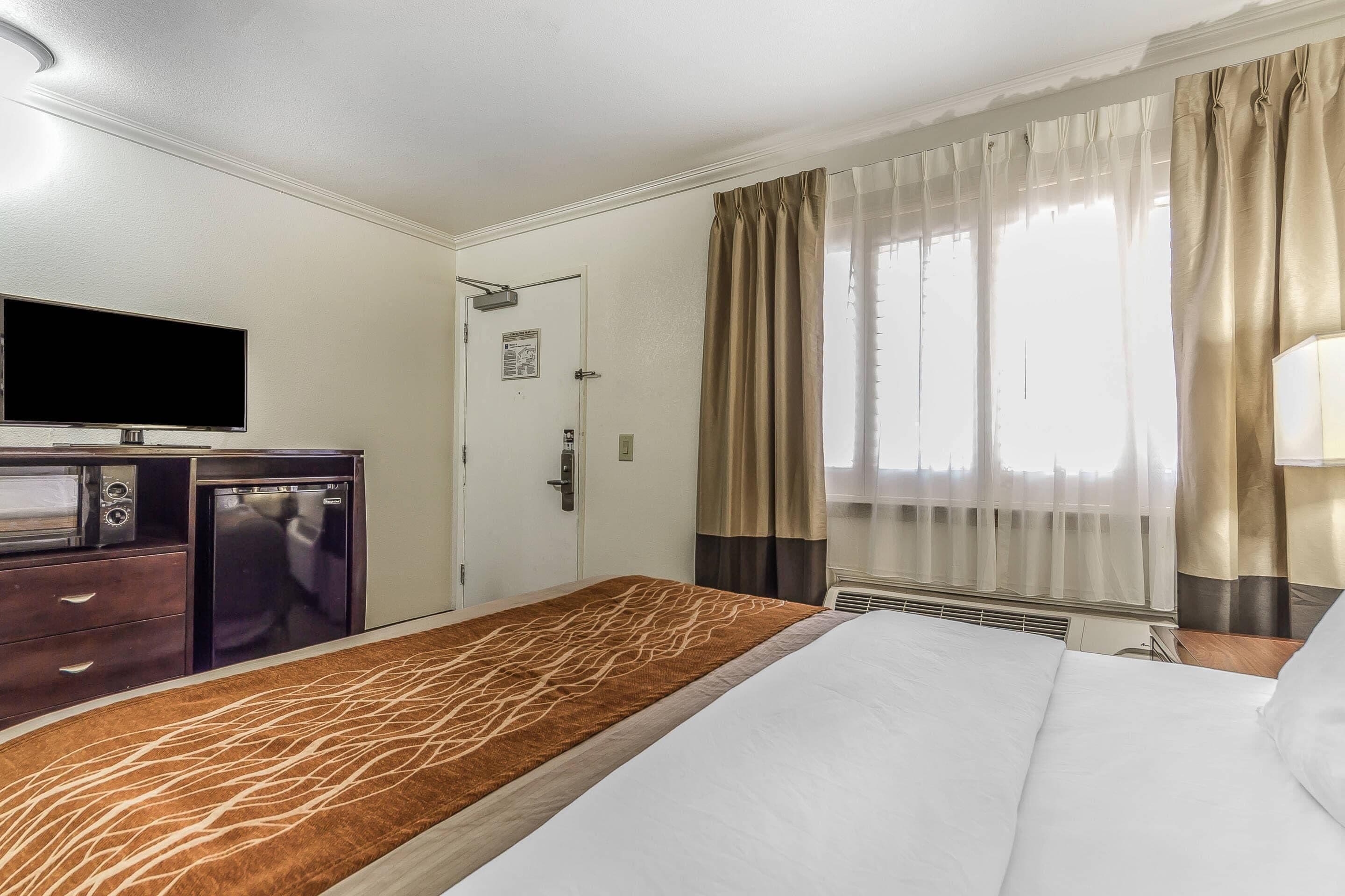 Habitación de hotel con cama, televisor de pantalla plana, mini refrigerador, microondas y escritorio con lámpara.