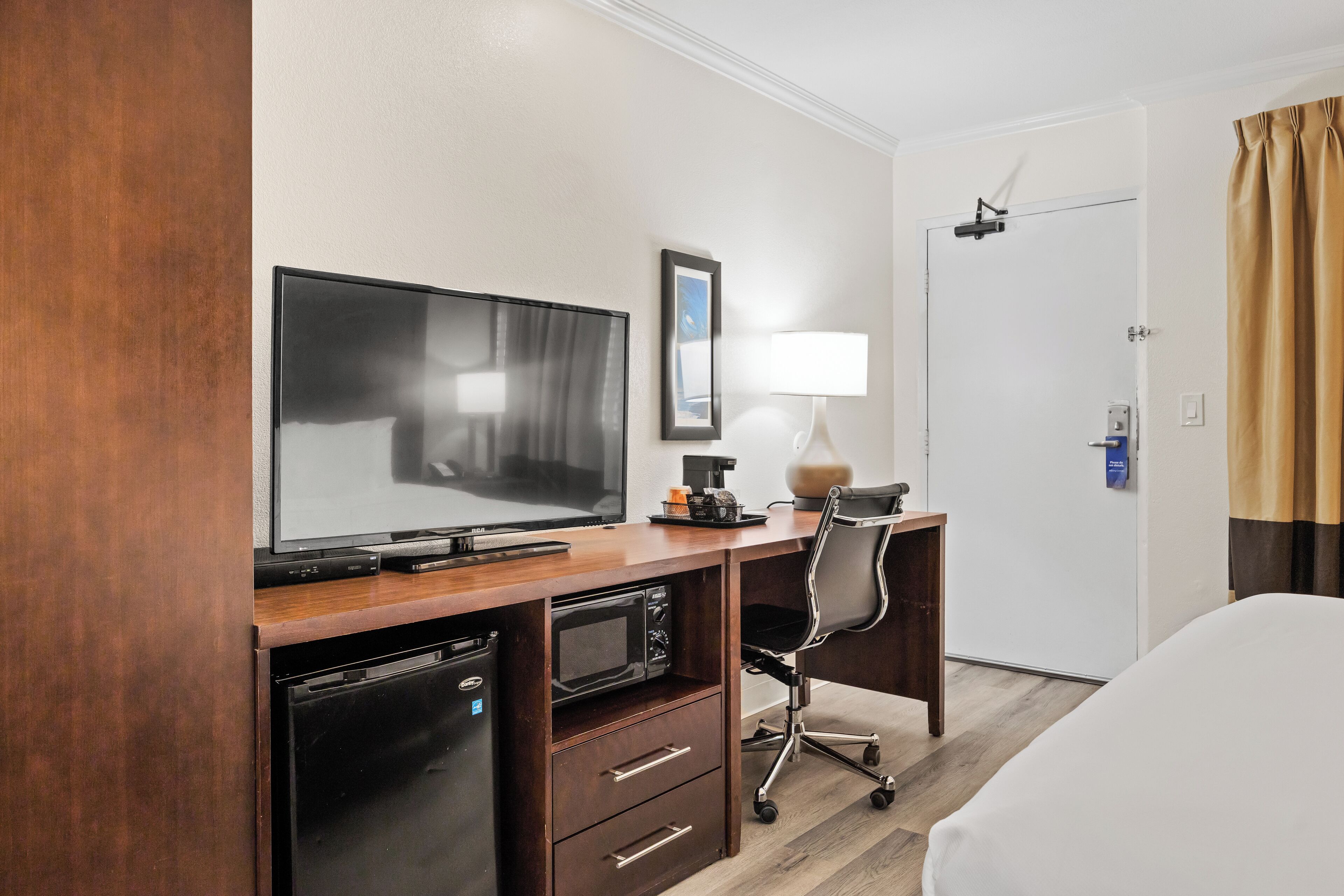 A hotel room with a wooden desk, a television, a microwave, a chair, a bed, a lamp, and a door with a curtain.