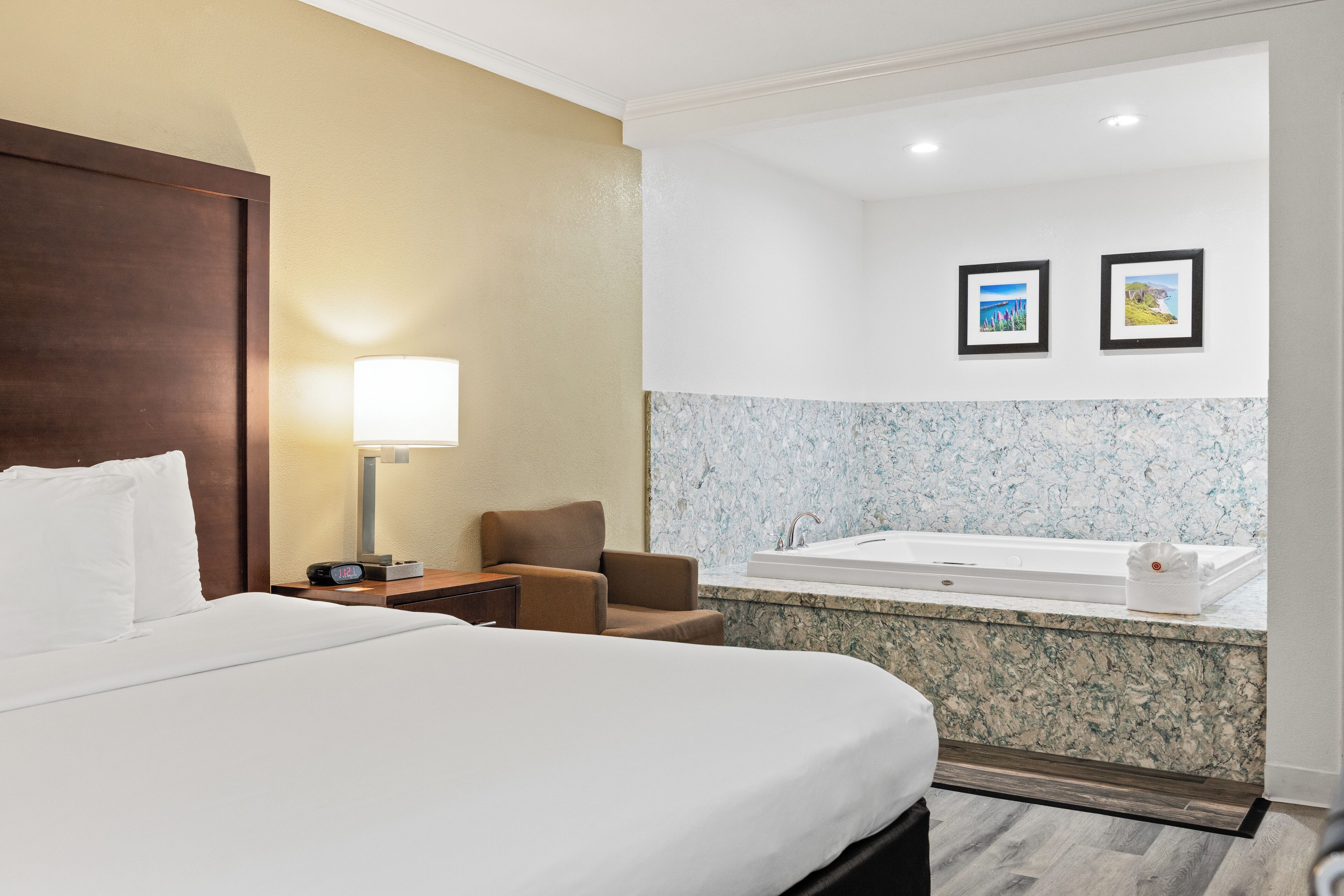 A hotel room with a large bed, a chair, a nightstand, a lamp, a bathroom with a bathtub, and two framed pictures on the wall.