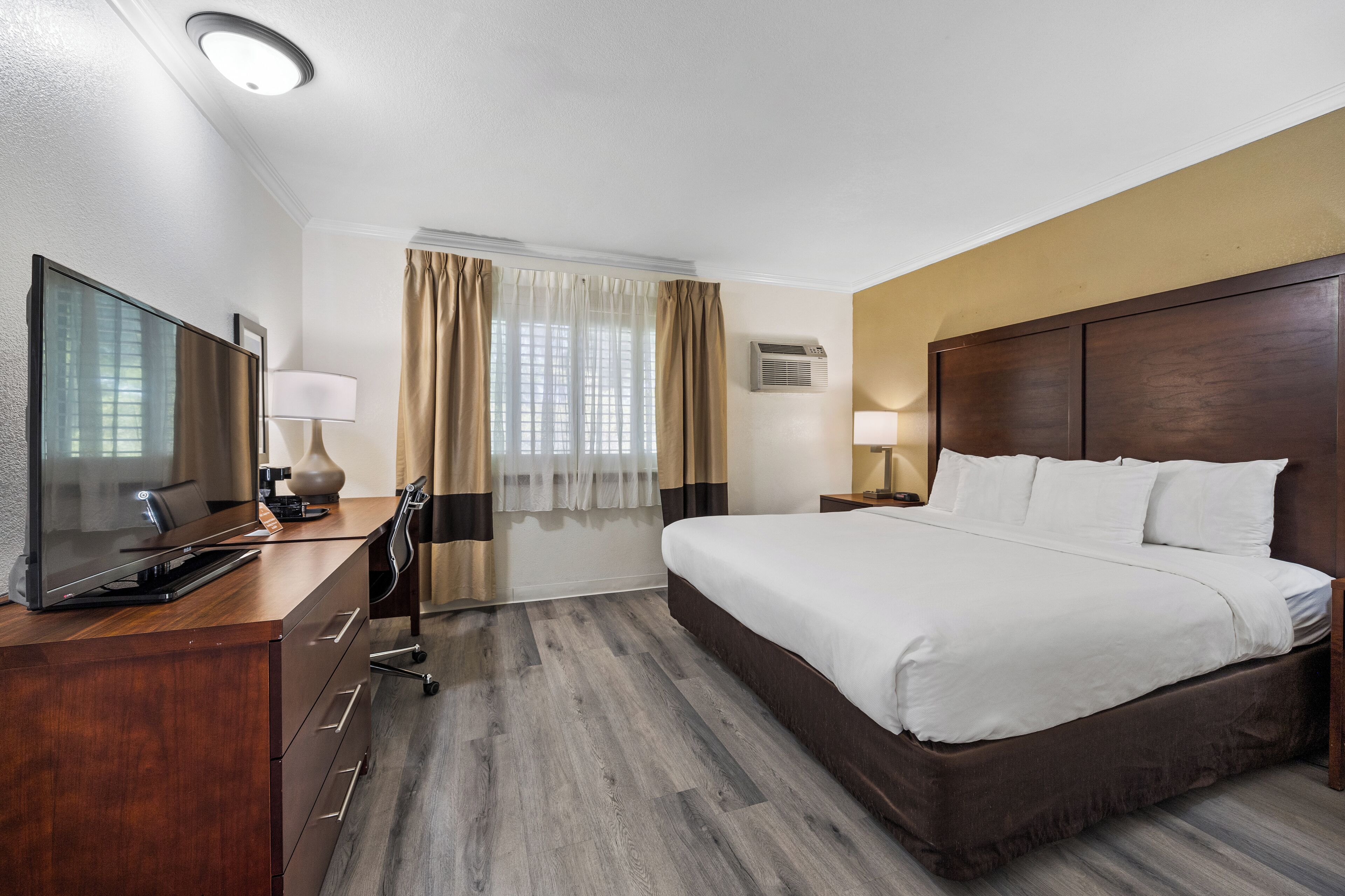 Ein Hotelzimmer mit einem großen Bett, einem Schreibtisch, einem Flachbildfernseher, einer Lampe und einem Fenster mit Vorhängen.