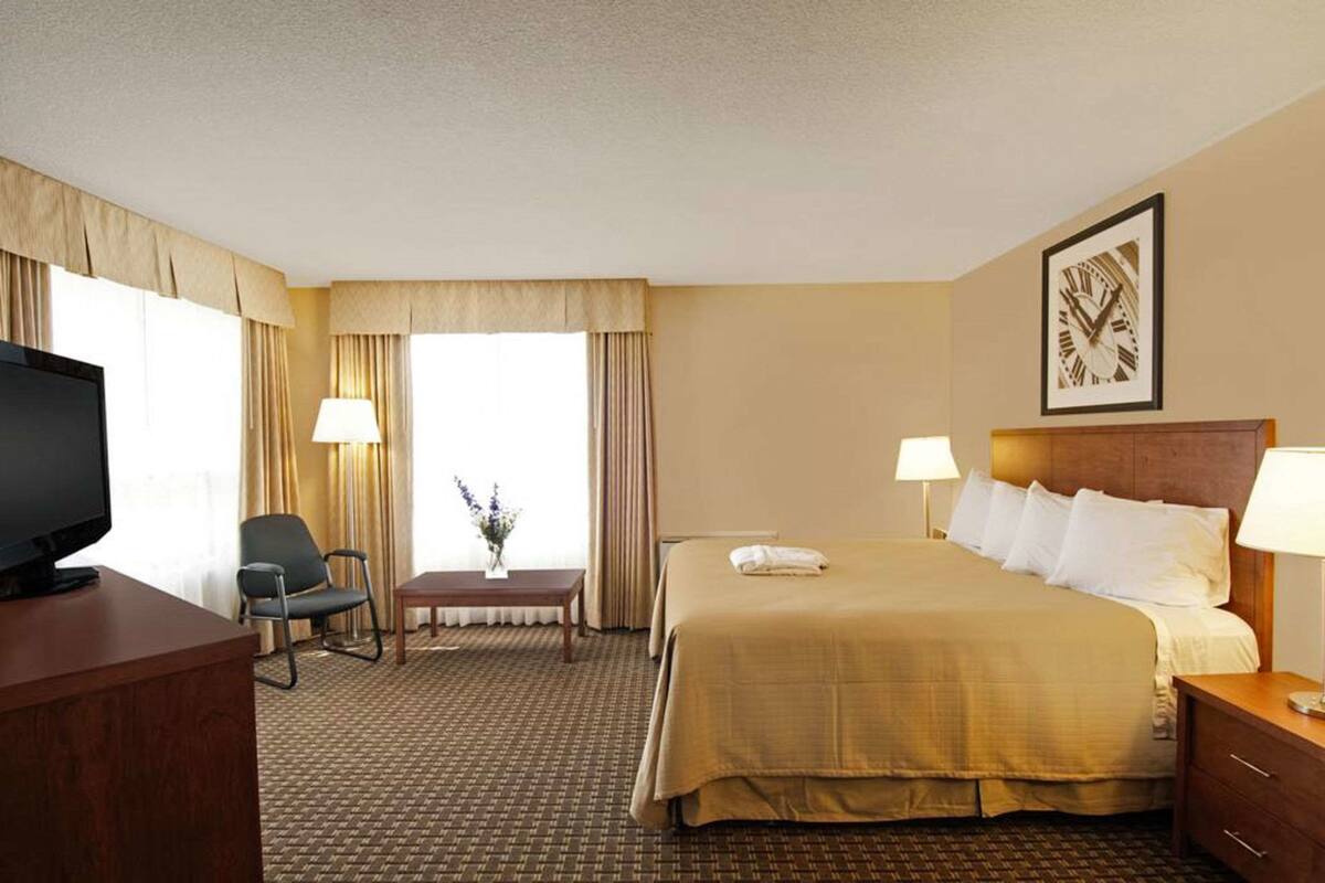A hotel room with a bed, bedside tables, a desk with a television, a chair, a small table with a vase, and a window with curtains.