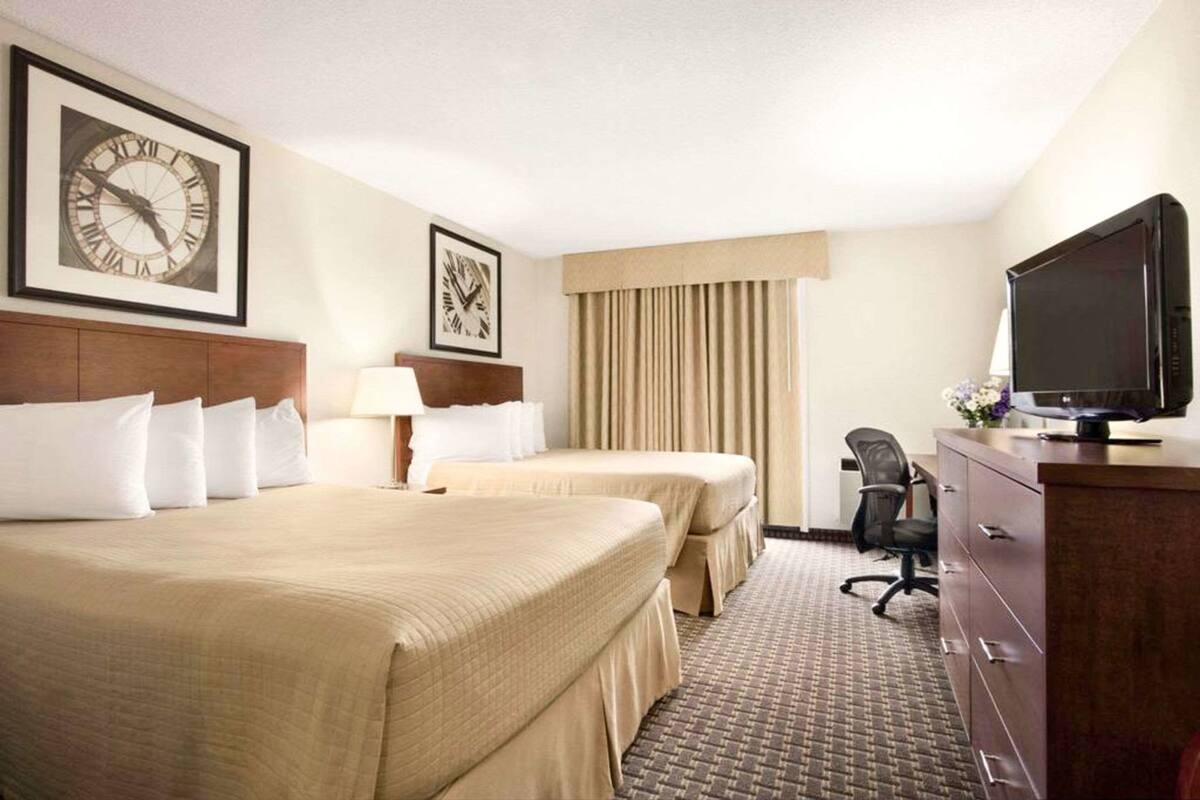 A hotel room with two beds, a television, a desk with a chair, and a clock on the wall.