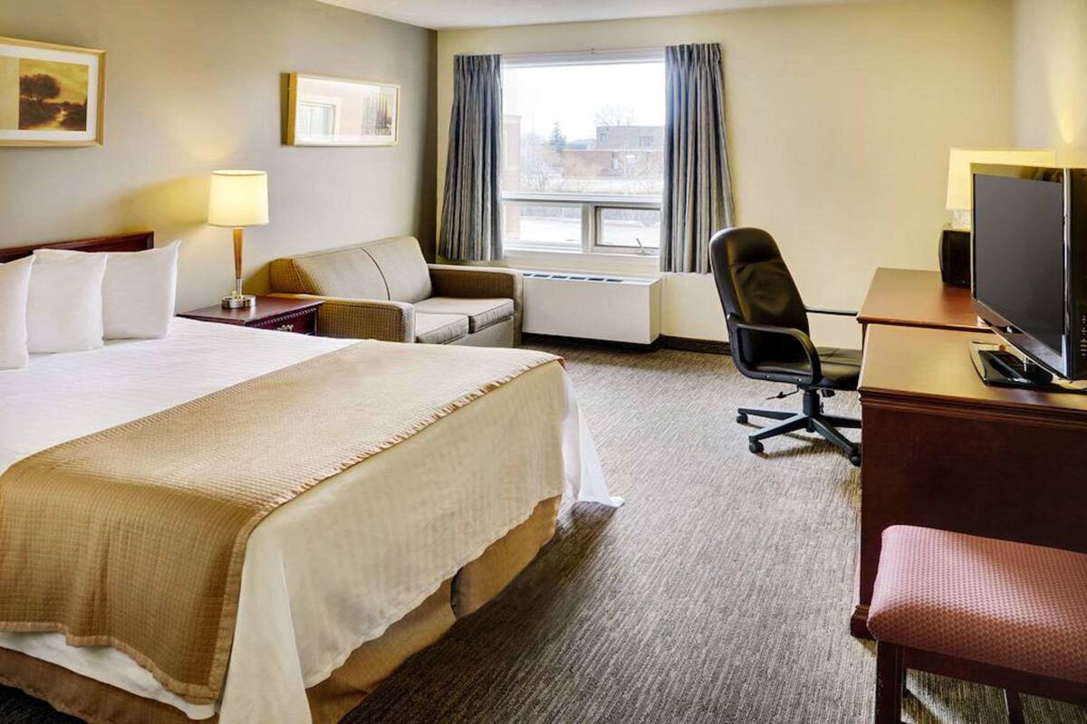 A hotel room with a large bed, a desk with a computer, a chair, a sofa, and a window with curtains.