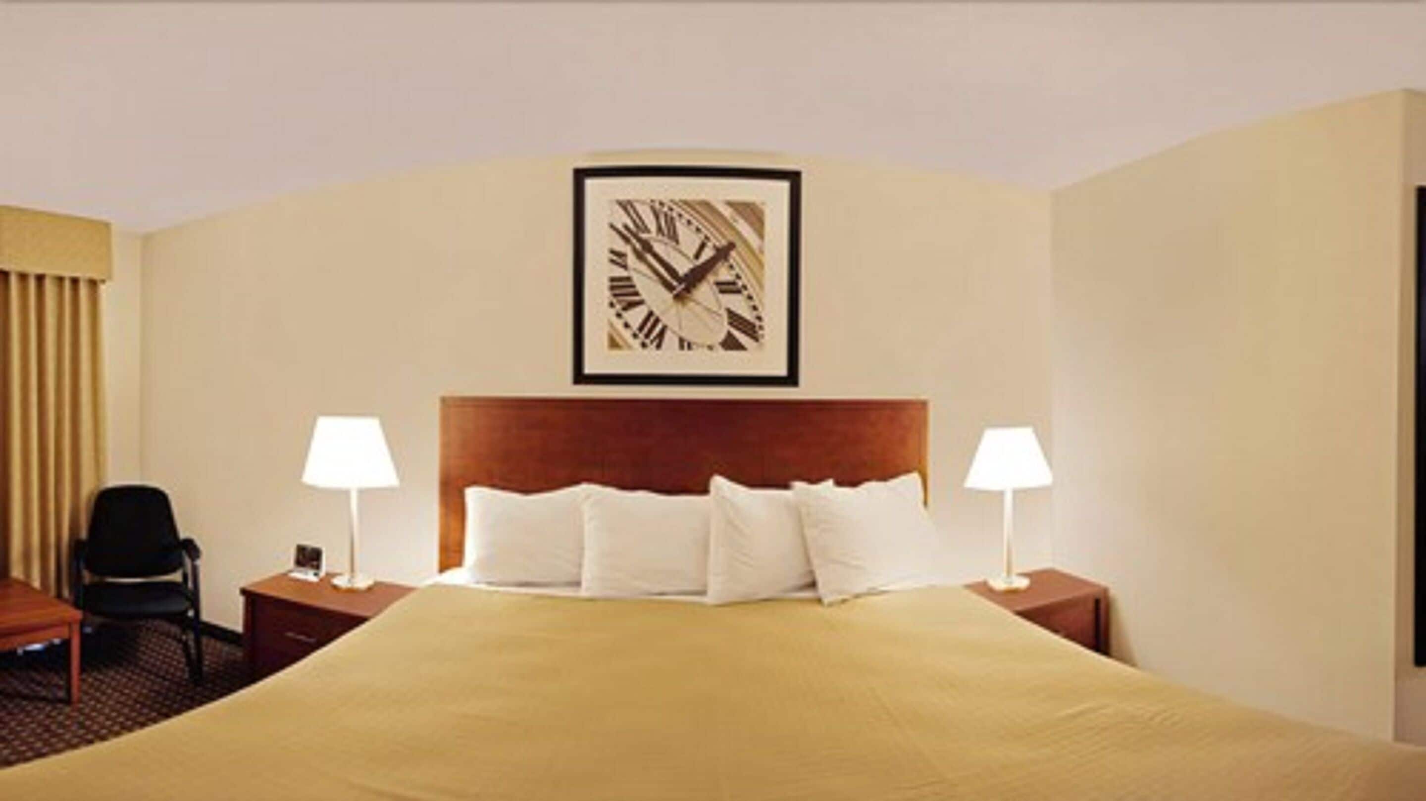 Habitación de hotel con una cama grande, dos mesitas de noche con lámparas, una silla y un reloj en la pared.