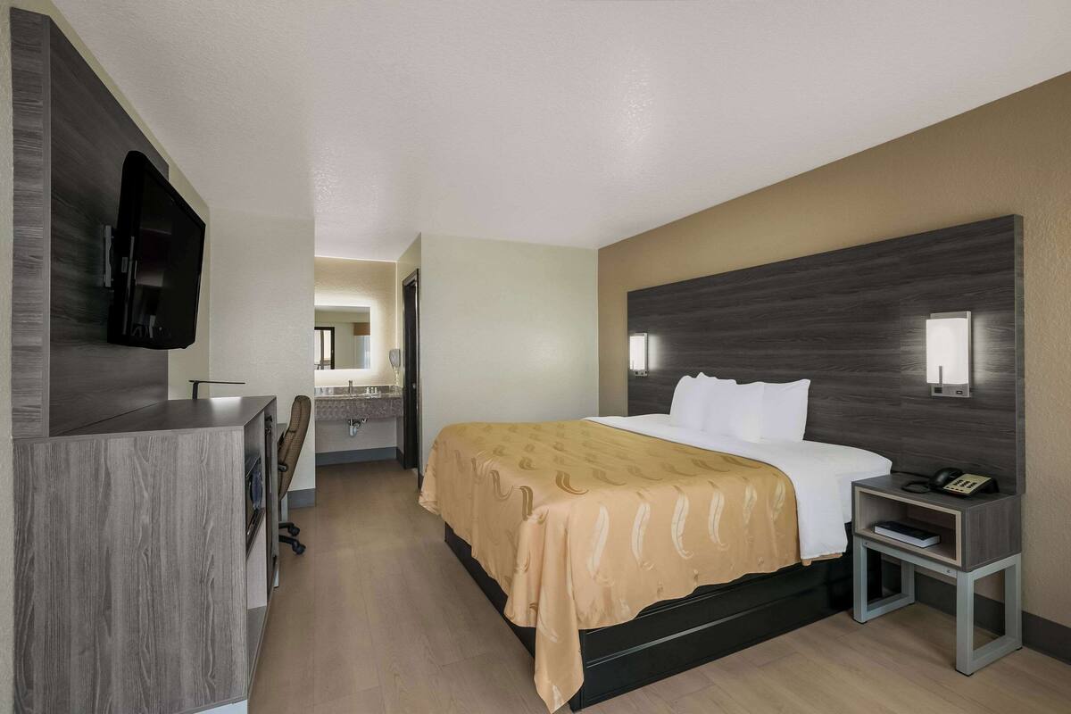 A hotel room with a bed, bedside table, TV, and a bathroom visible in the background.