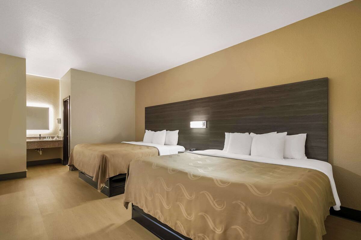 A hotel room with two beds, a large dark wood headboard, a bathroom with a mirror and sink, and a light-colored floor.